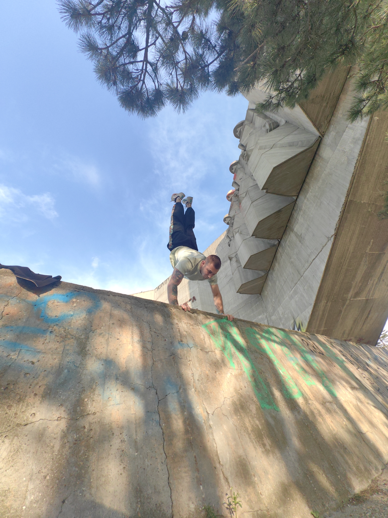 The image shows a man performing a handstand on the edge of a graffiti-covered concrete structure. The structure appears to be part of a larger concrete monument, possibly the Monument to the Bulgarian-Soviet Friendship in Varna, Bulgaria. The man is wearing a light-colored t-shirt and dark pants, and his feet are pointed upward. Above the man and the structure is a blue sky with some wispy clouds, partially obscured by tree branches. The photo is taken from a low angle, emphasizing the height of the structure and the man's position.