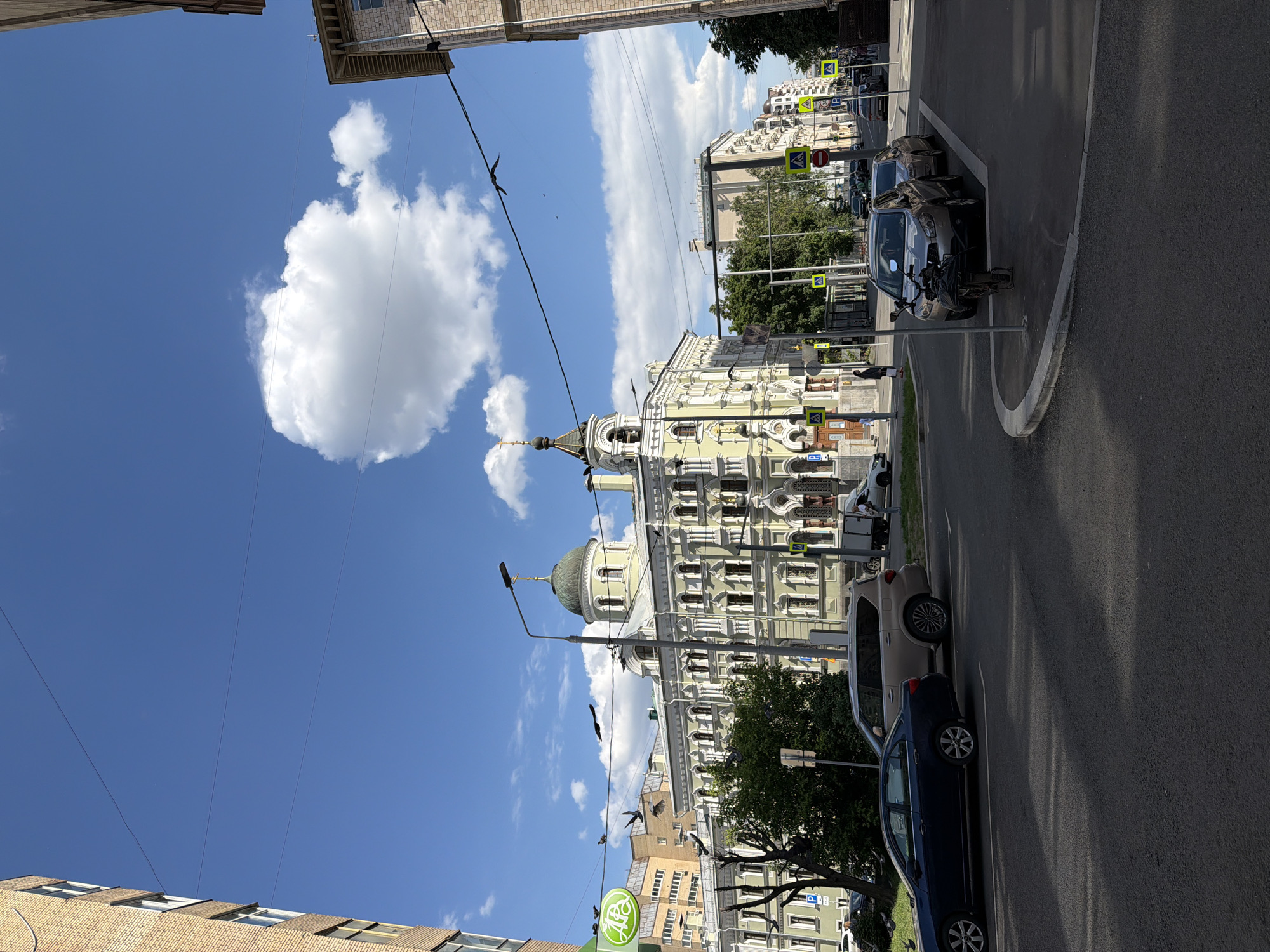 The image displays a sunny street scene in a city, likely Moscow, under a bright blue sky filled with scattered white cumulus clouds. The perspective looks down a street lined with buildings and vehicles.
On the right side of the street, a prominent, ornate building with light yellow-green walls and white decorative architectural elements captures attention. It features multiple windows, balconies, and a distinctive domed roof topped with a spire and a cross, suggesting it is a historical building or a church. Across the street, other buildings with varying facades, including a brick one, are visible.
The asphalt road, marked with pedestrian crossings, features several parked cars: a dark blue sedan, a beige SUV, and a silver car, alongside a motorcycle. A few individuals can be seen walking or standing in the distance. The street is equipped with typical urban infrastructure, including streetlights and overhead electrical wires, on which several birds are perched or flying.
Various road signs are visible, notably multiple yellow diamond-shaped pedestrian crossing signs and a red circular stop sign. A green circular sign with a white stylized Cyrillic "АГ" logo is visible near the bottom left, likely indicating a commercial establishment. The strong sunlight casts clear shadows, indicating it is midday or early afternoon. The scene conveys a sense of a peaceful yet active urban environment.