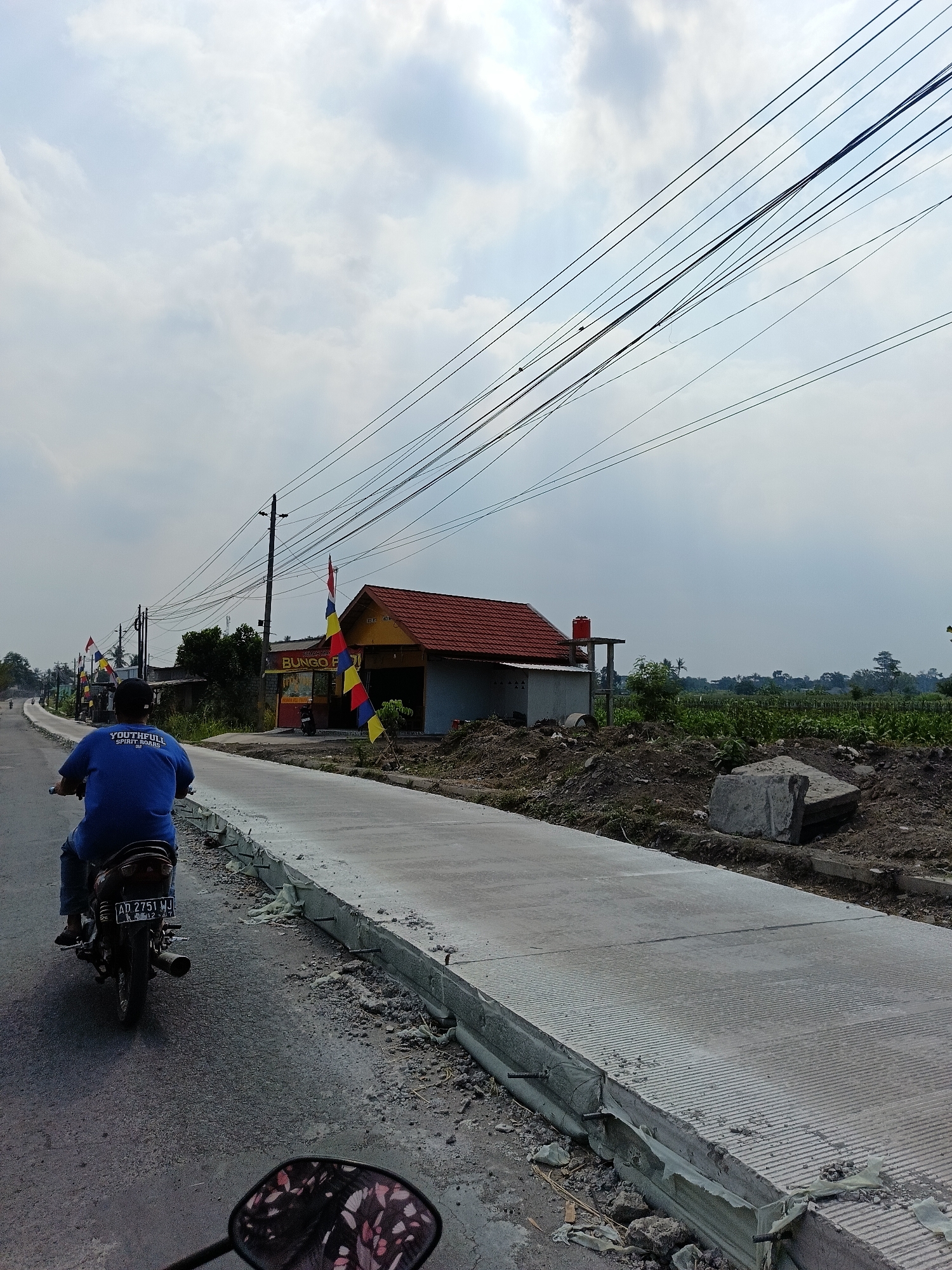 A motorcycle rider, wearing a blue shirt with white lettering, is traveling on a paved road in Manisrenggo, Indonesia. The rider is facing away from the camera. To the right of the road, a newly constructed concrete path is visible, with construction debris and raw materials scattered around. A small building with a red roof, possibly a shop or eatery, is situated next to the construction area, decorated with colorful flags. Overhead, a network of power lines stretches across the sky, which is partly cloudy. In the distance, a rural landscape with vegetation and trees can be seen. The license plate of the motorcycle is visible and reads "AD 2751 WJ".