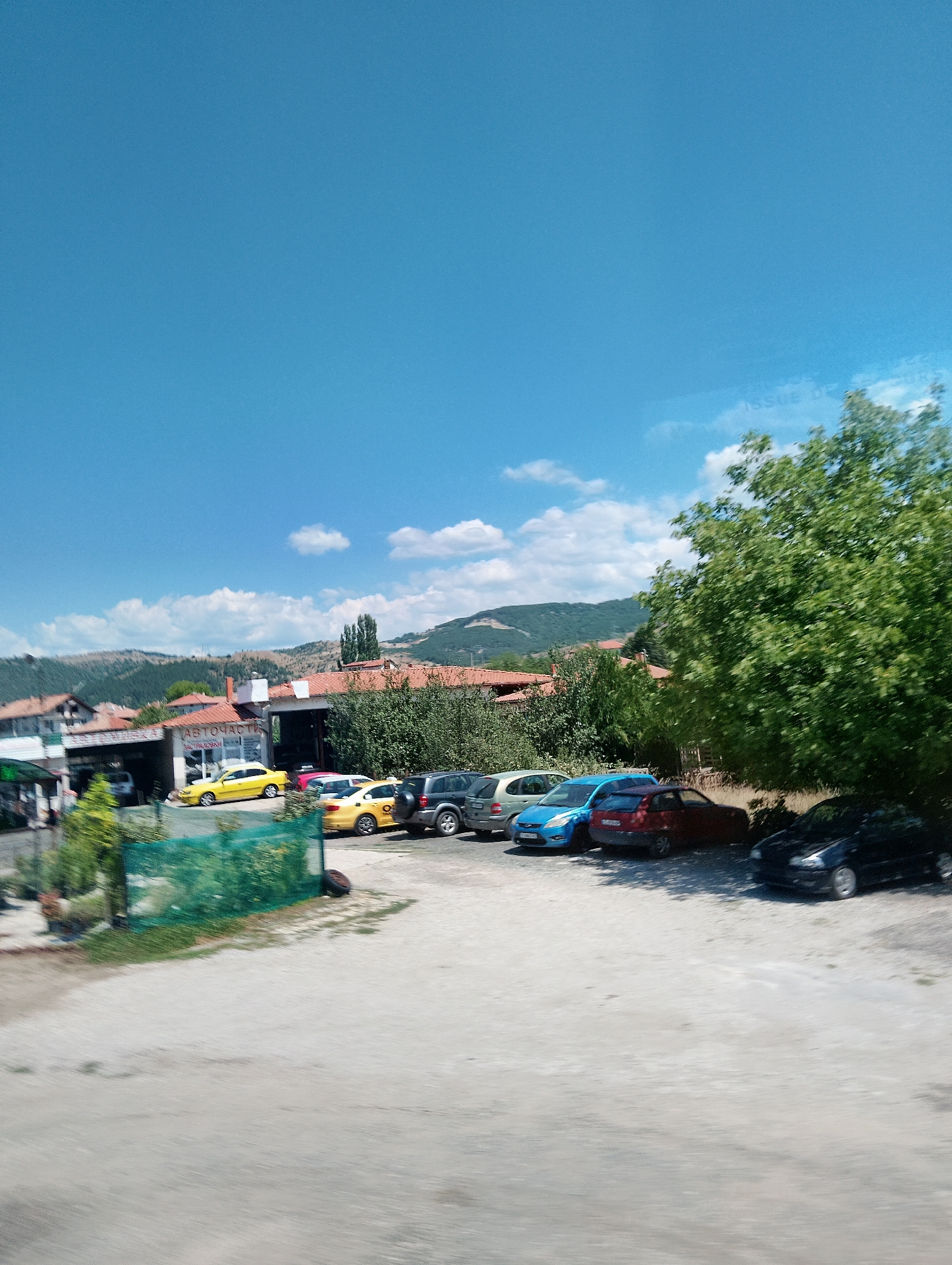 A bright, sunny day is depicted in Simitli, Bulgaria, with a clear blue sky dotted with scattered white clouds. The scene is a paved lot in front of a low-rise building with red tile roofs, possibly a business or service establishment. Several cars are parked in the lot, including a yellow taxi, a red car, a blue car, and a dark-colored SUV. A large, leafy green tree stands to the right of the frame, casting a shadow over some of the parked vehicles. The background shows rolling hills covered with vegetation, indicative of the Bulgarian landscape. There are no people visible in the image. The overall impression is one of a quiet, possibly rural, commercial area.