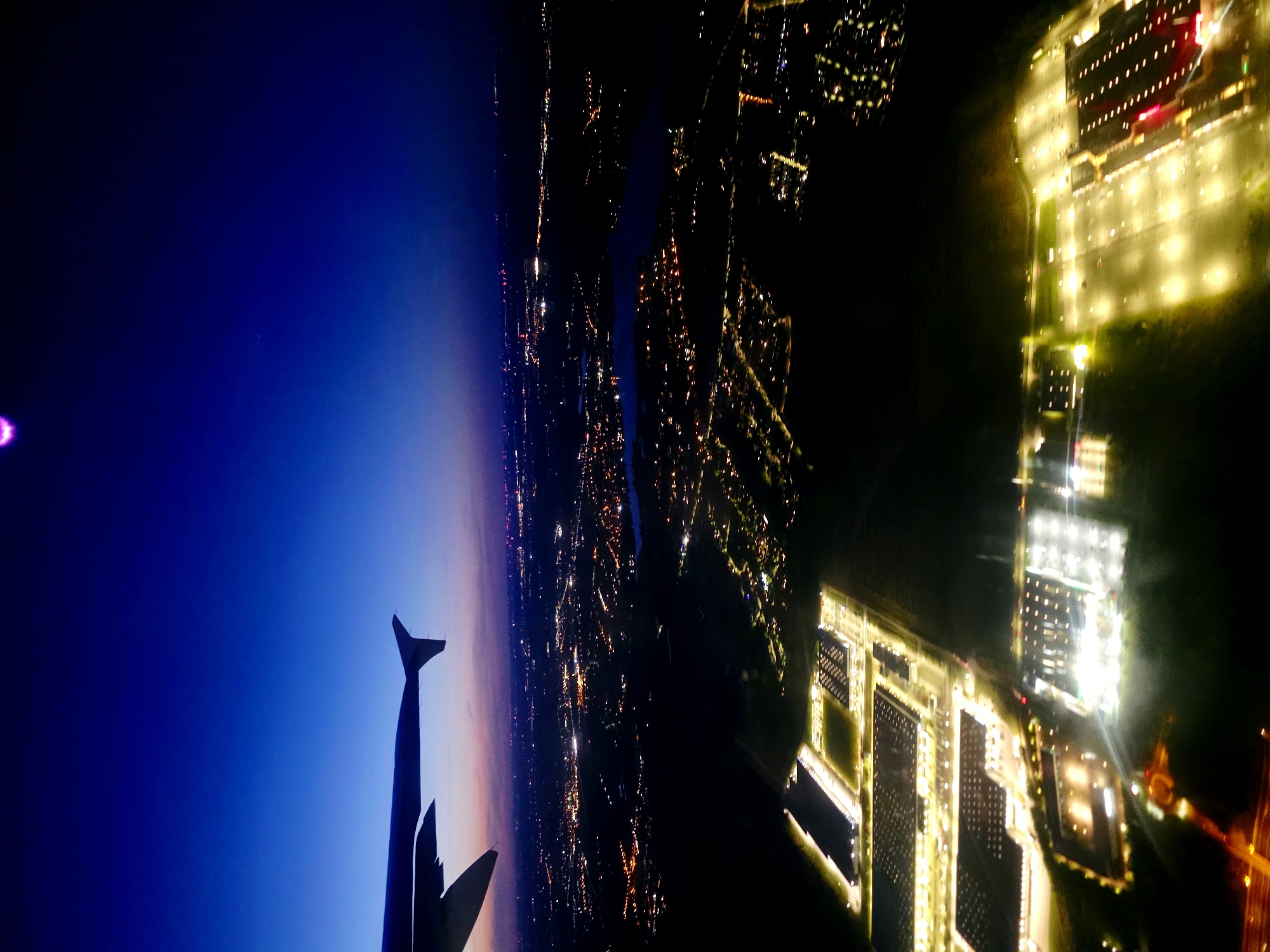 The image, taken from an airplane at dusk, shows an aerial view of the city of Swadzim, Poland. The sky transitions from a deep blue overhead to hues of pink and orange near the horizon. The wing of the airplane is visible in the upper left corner. Below, a sprawling urban landscape is illuminated by a network of city lights, indicative of residential and commercial areas. In the foreground, a large industrial complex with multiple warehouses is brightly lit, with rows of cars visible in its parking lots. A body of water, possibly a lake, is visible in the middle ground, reflecting the city lights. The overall scene depicts a vibrant, illuminated city at twilight.