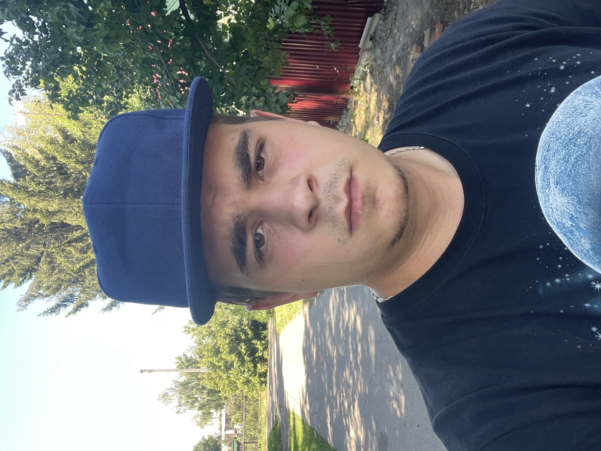 A young man wearing a dark blue flat-brimmed baseball cap and a black t-shirt stands outside on a paved path in Joğarğy Berezovka, Kazakhstan. The man has brown eyes and a light complexion, with a small goatee. He is looking directly at the camera, with a neutral expression. Behind him, the path continues into the distance, bordered by grass and trees. A wooden fence can be seen on the left side of the path, and a red metal fence is visible on the right. The background is filled with lush green foliage and tall trees. The sun is shining brightly, casting shadows on the path. The overall impression is a casual outdoor portrait taken on a sunny day in a rural or semi-rural setting.