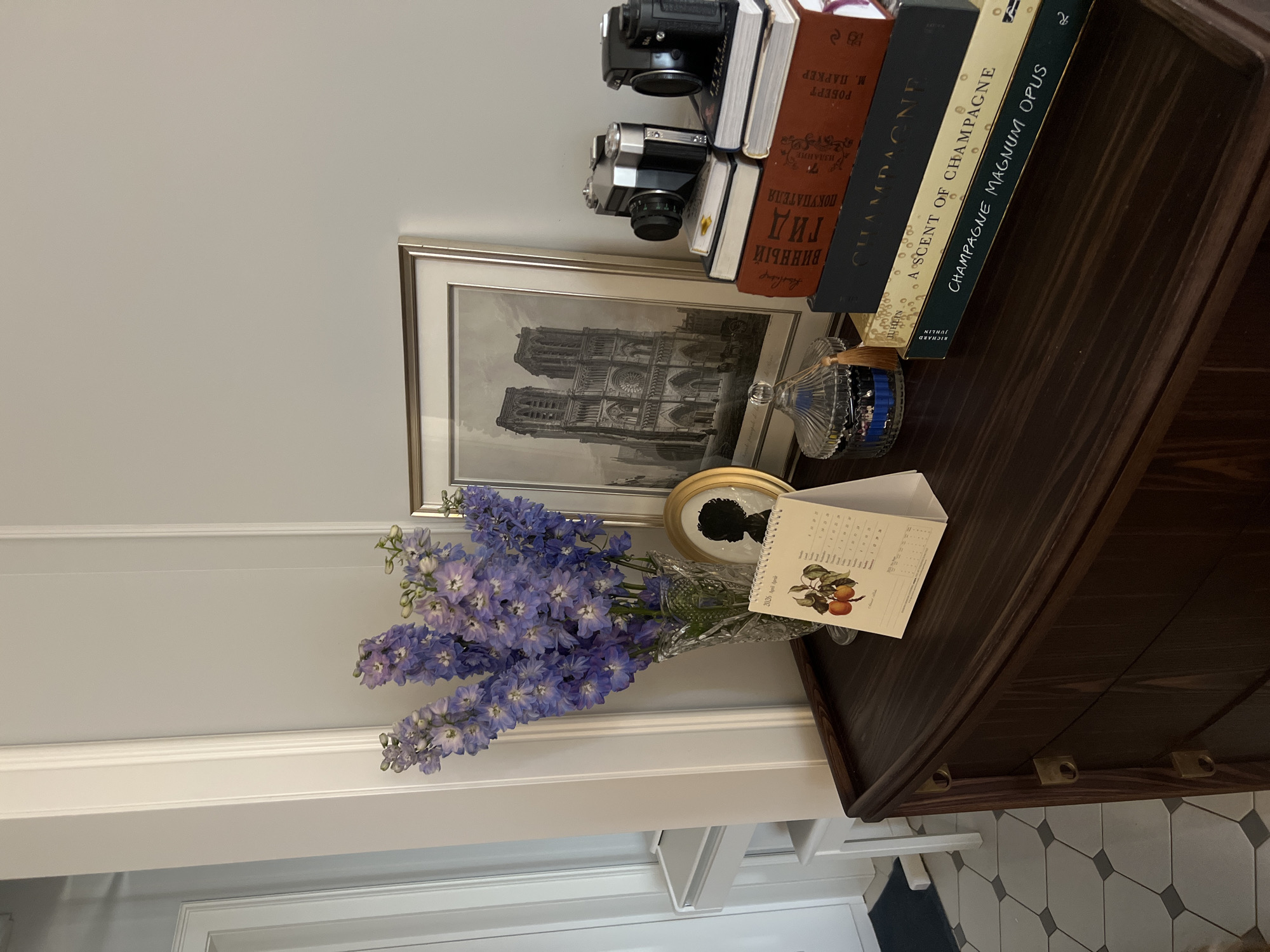 This media file captures a decorative still life arrangement on a dark wooden surface, likely a cabinet or console table, set against a light-colored wall. A prominent bouquet of vibrant purple delphinium flowers, in full bloom, is displayed in a clear, ornate glass vase on the left. Behind the flowers, a framed black-and-white print of Notre Dame Cathedral is visible. To its right, a small gold-framed silhouette portrait and a desk calendar open to "2026 April" with a fruit illustration are arranged.
Above the framed picture, several books are stacked. Two vintage cameras, one black and one silver, rest atop a horizontal stack of books. Visible book titles include "A SCENT OF CHAMPAGNE," "CHAMPAGNE MAGNUM OPUS," and a partially visible "CHAMPAGNE." Other books bear Cyrillic text, including "РОБЕРТ М. ПАРКЕР" (Robert M. Parker), "ТИД", "ВИННЫЙ", "ПОКУПАТЕЛЯ", and "ИЗДАНИЕ," indicating a connection to Russia. A small, clear glass bottle with a blue stopper, possibly an inkwell, is also part of the display. The wall features subtle paneling detail, and the floor visible at the bottom right has a geometric grey and off-white tile pattern. The scene is static, showcasing a carefully curated collection of objects. The Cyrillic text on the books supports the given context of Moscow, Russia.
