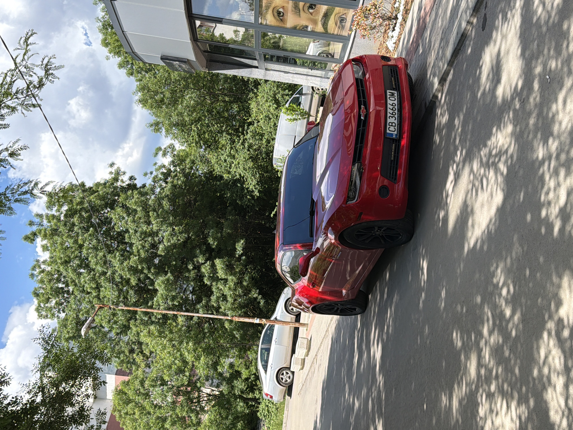The image depicts a red Chevrolet Camaro parked on a concrete pavement, with the front of the vehicle facing right. The car has a license plate that reads "CB 3666 OM". Behind the Camaro, there is a white sedan and a white van, all parked along the curb. The background includes a building with reflective windows and a large green tree providing shade over the scene. A utility pole is visible near the left side of the image. The setting appears to be an urban street in Unknown, Unknown on a sunny day. The reflection of eyes is visible in the building window.
