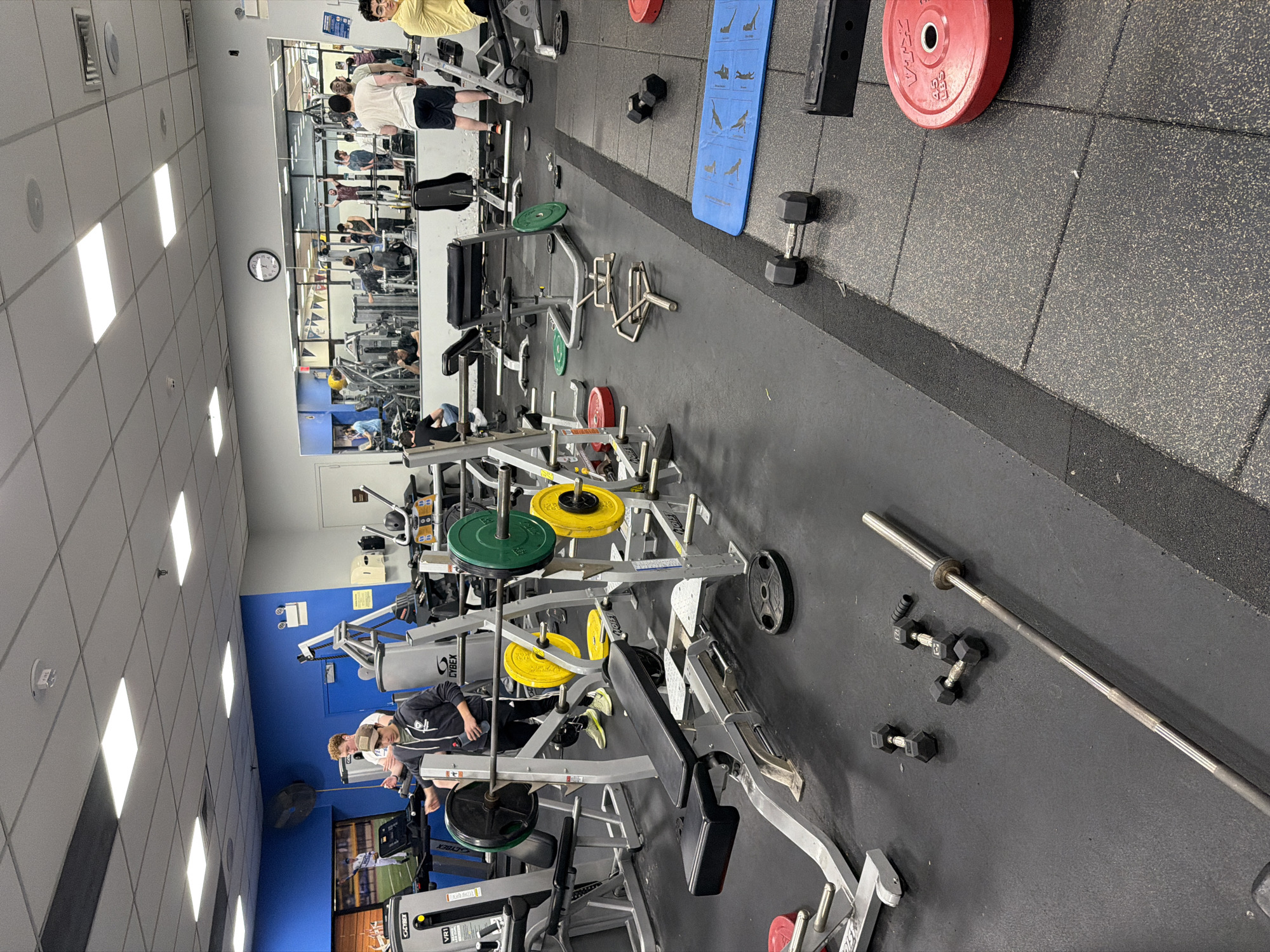 This image depicts the interior of a gym, likely in the city of New York, United States of America. The gym is filled with various weightlifting machines and equipment, including leg press machines, benches, and dumbbells. There are several people present, some of whom appear to be working out. The environment is typical of a commercial gym, with rubber flooring, mirrored walls, and fluorescent lighting.
