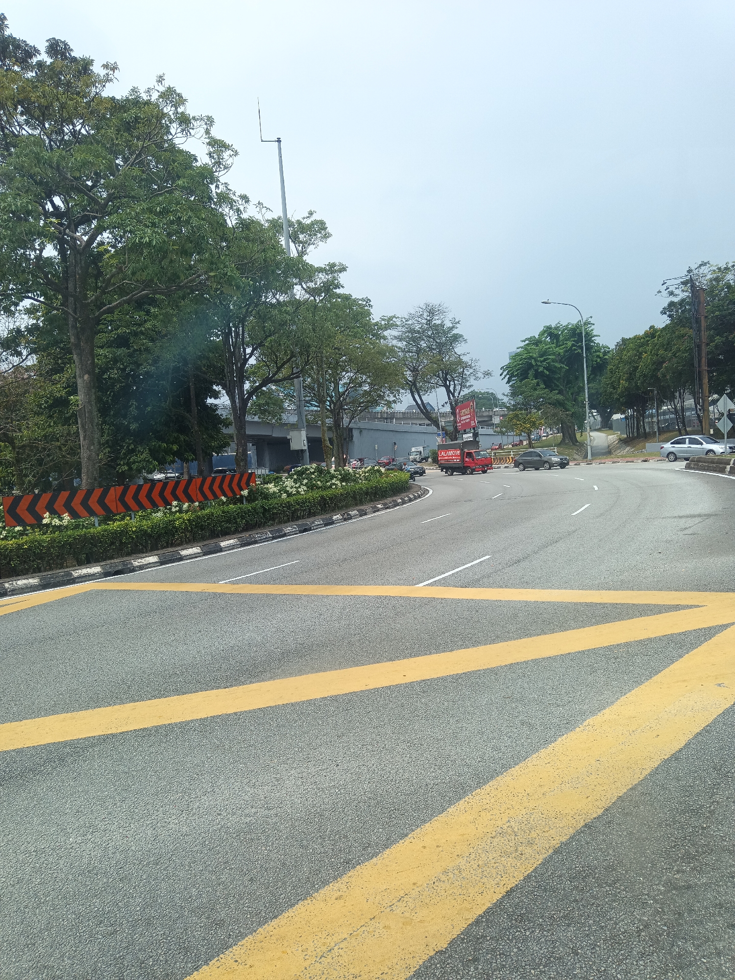 This is a photograph taken at the intersection of Jalan Tuanku Abdul Halim and Jalan Damansara in Kuala Lumpur, Malaysia. The viewpoint is from a road with prominent yellow directional arrows painted on the asphalt. The road curves to the right, leading towards a traffic light. On the left side of the road, there is a chevron-patterned traffic barrier with black and orange markings, separating the lanes from a green median strip with manicured bushes and some white flowers. Mature trees with dense green foliage are visible on the left and in the background. In the distance, an elevated highway structure is present. Several vehicles are on the road, including a red Lalamove delivery truck, a dark gray sedan, and a white sedan. The sky is overcast, suggesting a cloudy day. The environment appears to be a busy urban road network.