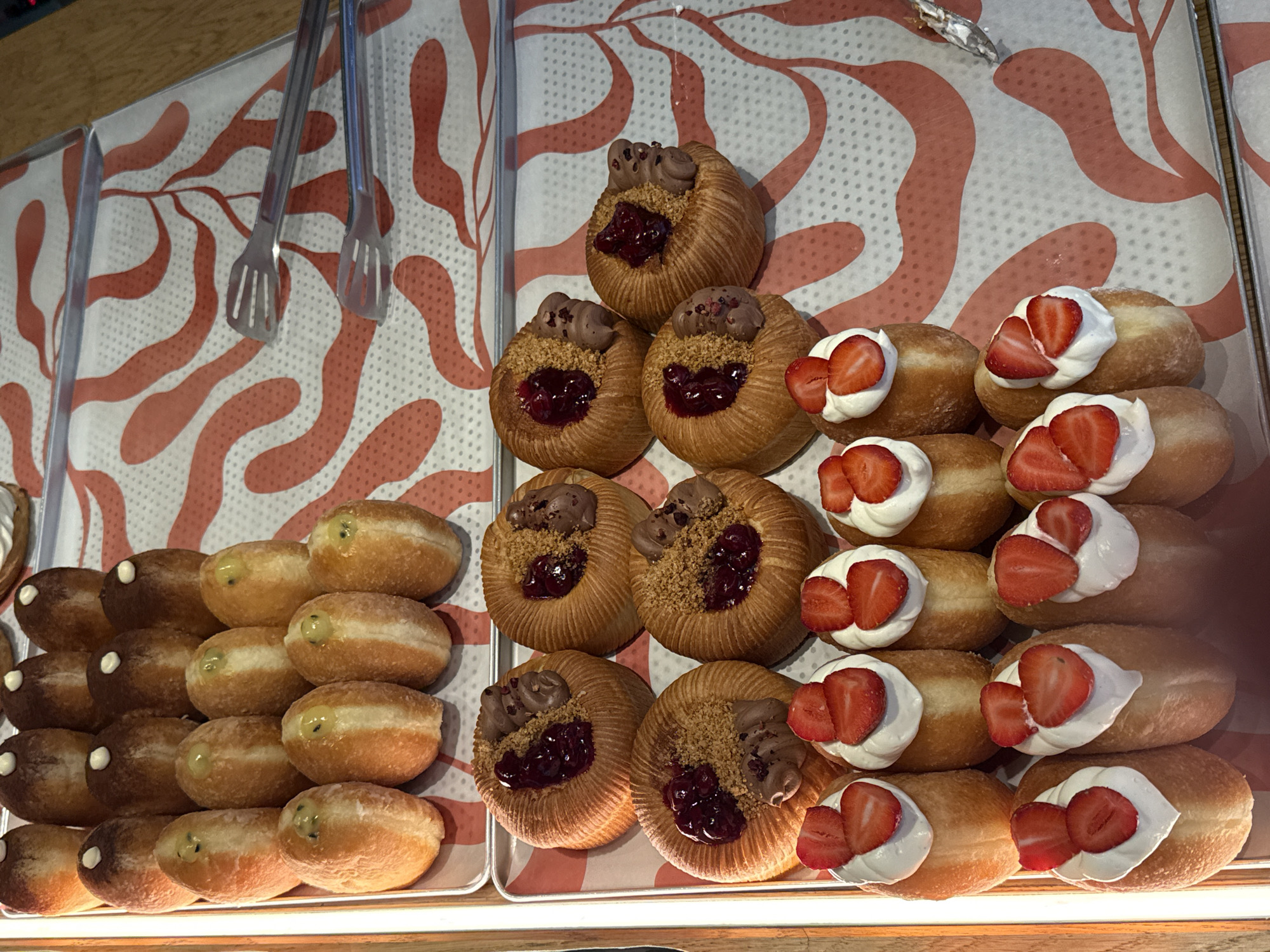 This media file shows a display of pastries, likely in a bakery or cafe. The pastries are arranged on trays with a decorative pattern of pink, abstract shapes on a white background. Two pairs of metal tongs are visible at the top left, suggesting self-service or preparation for serving.
The subjects are primarily the pastries themselves. On the left, there are several ring-shaped pastries with a golden-brown, flaky exterior, filled with a creamy filling that appears light green, possibly pistachio or lime flavored. Some of these also have a dark brown topping. In the center, there are rounded pastries with a similar golden-brown crust, topped with chocolate frosting, a dollop of red jam or fruit filling, and sprinkled with what looks like crushed nuts or cookie crumbs. To the right, a row of oblong-shaped pastries are filled and topped with whipped cream and fresh strawberry slices.
The setting appears to be indoors, given the lighting and the presence of a wooden surface at the top edge of the frame. The overall impression is one of abundance and tempting treats, ready to be purchased and enjoyed. There is no visible text in the image. The bright lighting suggests it could be daytime. The pastries are fresh and appealing, implying a commercial environment focused on baked goods. The context of Saint Petersburg, Russia, suggests this could be a local bakery offering a variety of sweet items.
