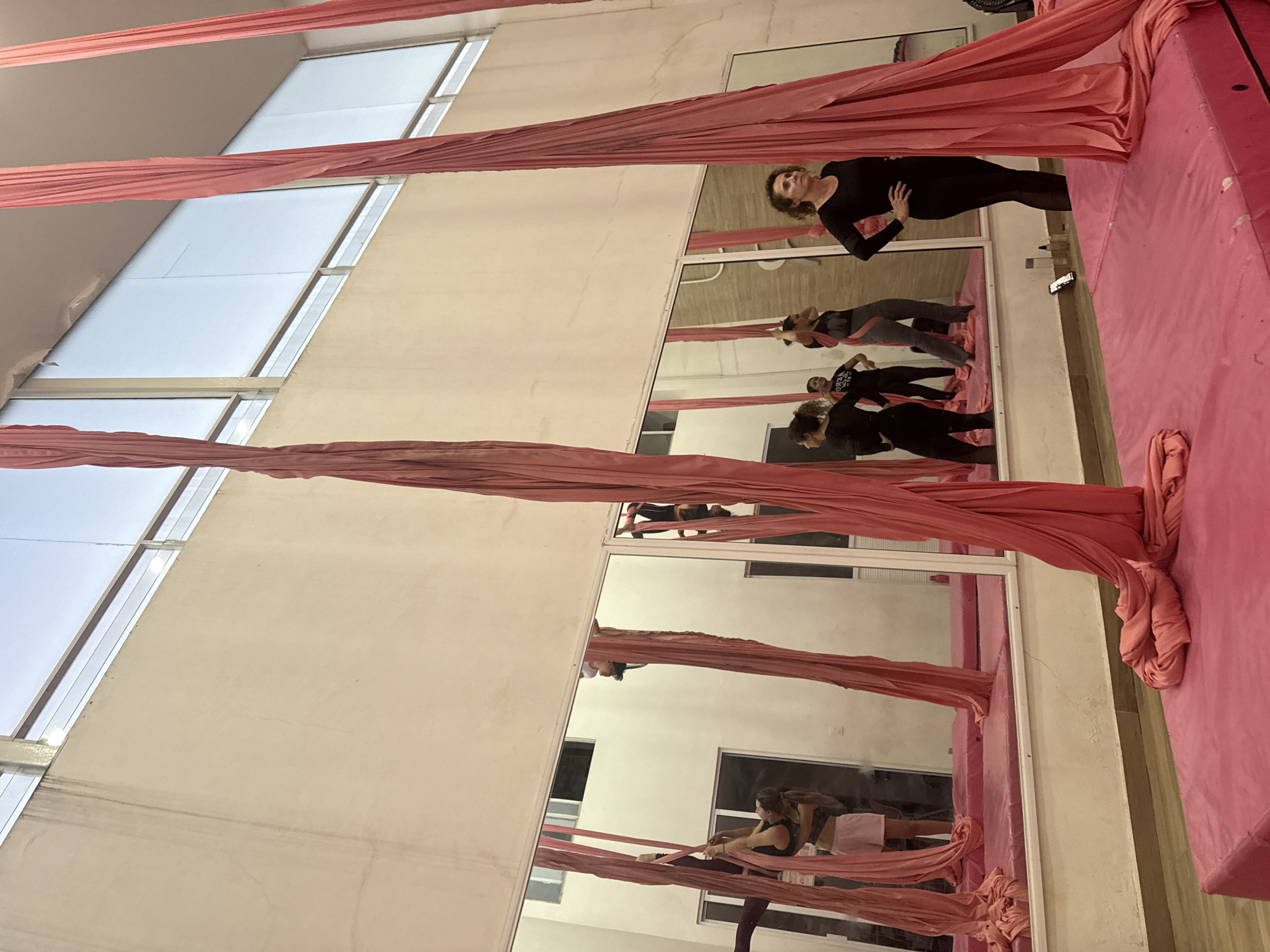 The image depicts an indoor aerial silks studio, likely located in San Andrés Cholula, Mexico, characterized by its high ceilings, large mirrors, and specialized equipment. The scene appears to take place during the daytime, with natural light entering through long, rectangular windows positioned high on the wall.
The primary subjects are several women engaged in an aerial silks practice or class. In the foreground, a woman dressed in black athletic attire stands on a pink safety mat, posing or observing, with her gaze directed upwards. Her reflection, along with those of other participants, is prominently displayed in the large mirrors that line the studio wall. These reflections show other women in various stages of activity: one woman in a grey top and dark pants stands with her hands on her hips, another wears a dark top with the partial text "VERO" visible on it, and a third woman in dark clothing appears to be stretching or interacting with the pink silks, which are draped around her leg.
The studio's setting includes light-colored walls, pink safety mats covering the floor, and multiple strands of vibrant pink aerial silks hanging from the ceiling, ready for use. A small, dark object, possibly a smartphone, is visible resting on one of the pink mats. The overall atmosphere suggests a focused and dedicated environment for physical training in aerial arts.