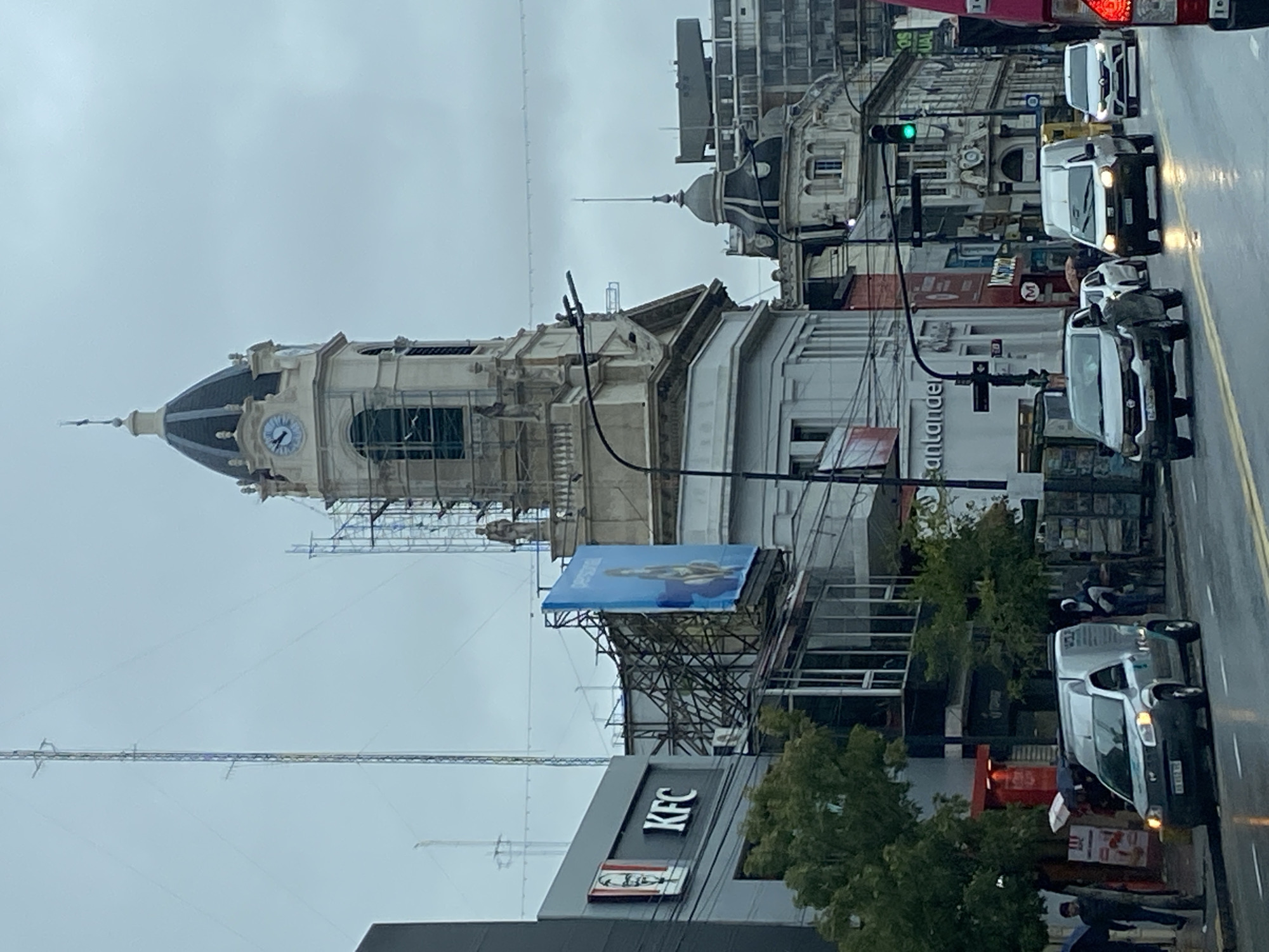The image shows a street scene in Buenos Aires, Argentina. In the foreground, a KFC restaurant is visible, partially obscured by trees. Further back, a large, white building with a clock tower is under construction, with scaffolding around it. A blue billboard is attached to a building near the clock tower. The street is wet from recent rain, and traffic is visible, including several cars and vans. Santander bank is identifiable on a building across the street. Pedestrians are on the sidewalks. The sky is overcast, indicating a cloudy day.