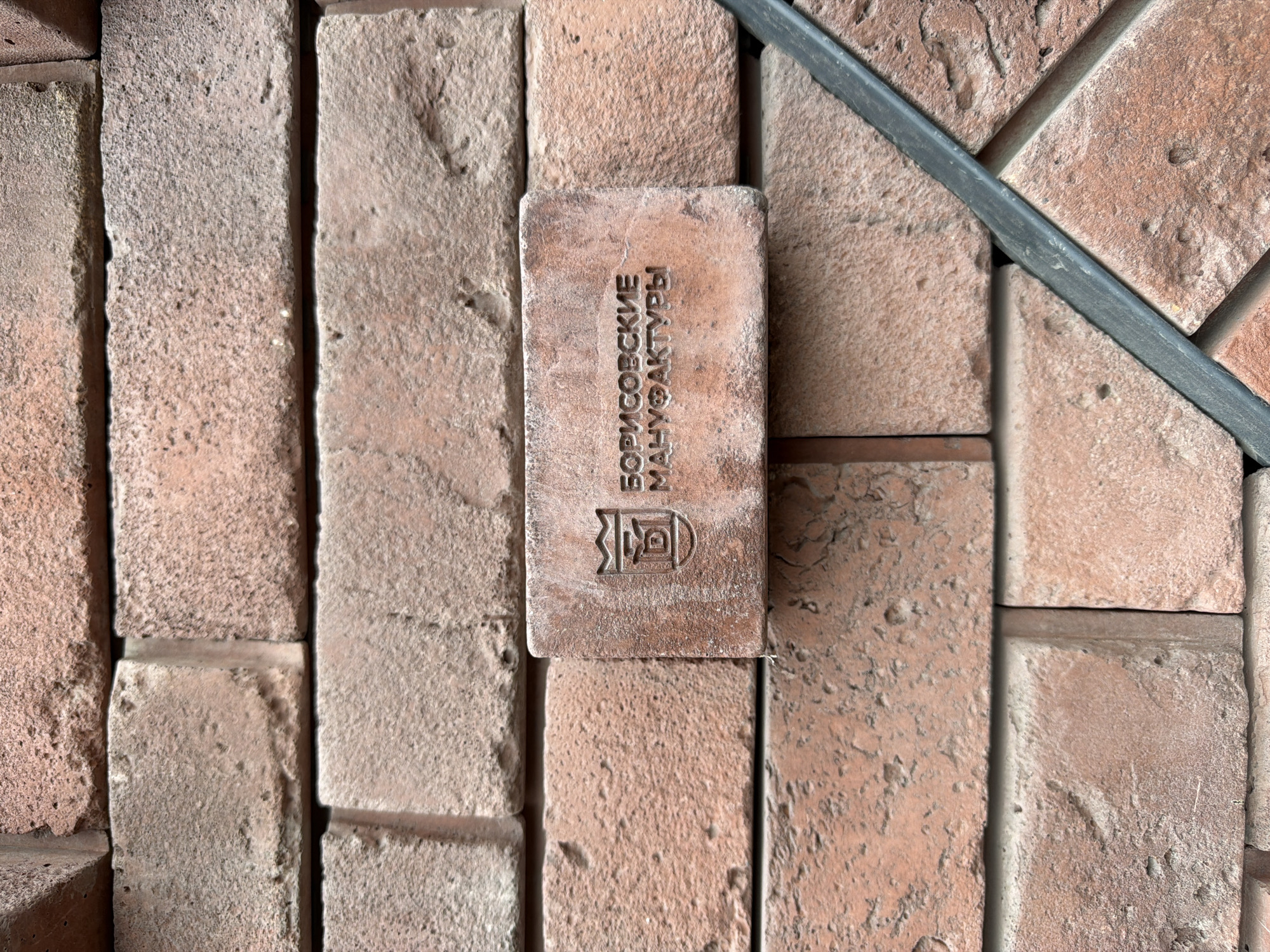 The image shows a close-up view of a weathered brick wall in Saint Petersburg, Russia. A single brick with an embossed logo and text is prominently featured in the center. The logo appears to be a shield with a crown on top and a stylized depiction of a building or crest inside. The text below the logo reads "БОРИСОВСКИЕ МАНУФАКТУРЫ," which translates to "Borisov Manufactories." The bricks are a reddish-brown color and show signs of age and wear, with some chipped edges and rough textures. A dark, thin, flexible material, possibly a sealant or trim, runs diagonally across the lower right portion of the frame, nestled between bricks. The background consists of more bricks, arranged in a traditional pattern. There are no people or animals visible in the image. The lighting suggests it is daytime, with some shadows indicating the direction of the light source. The overall impression is one of a rustic or historical architectural detail.