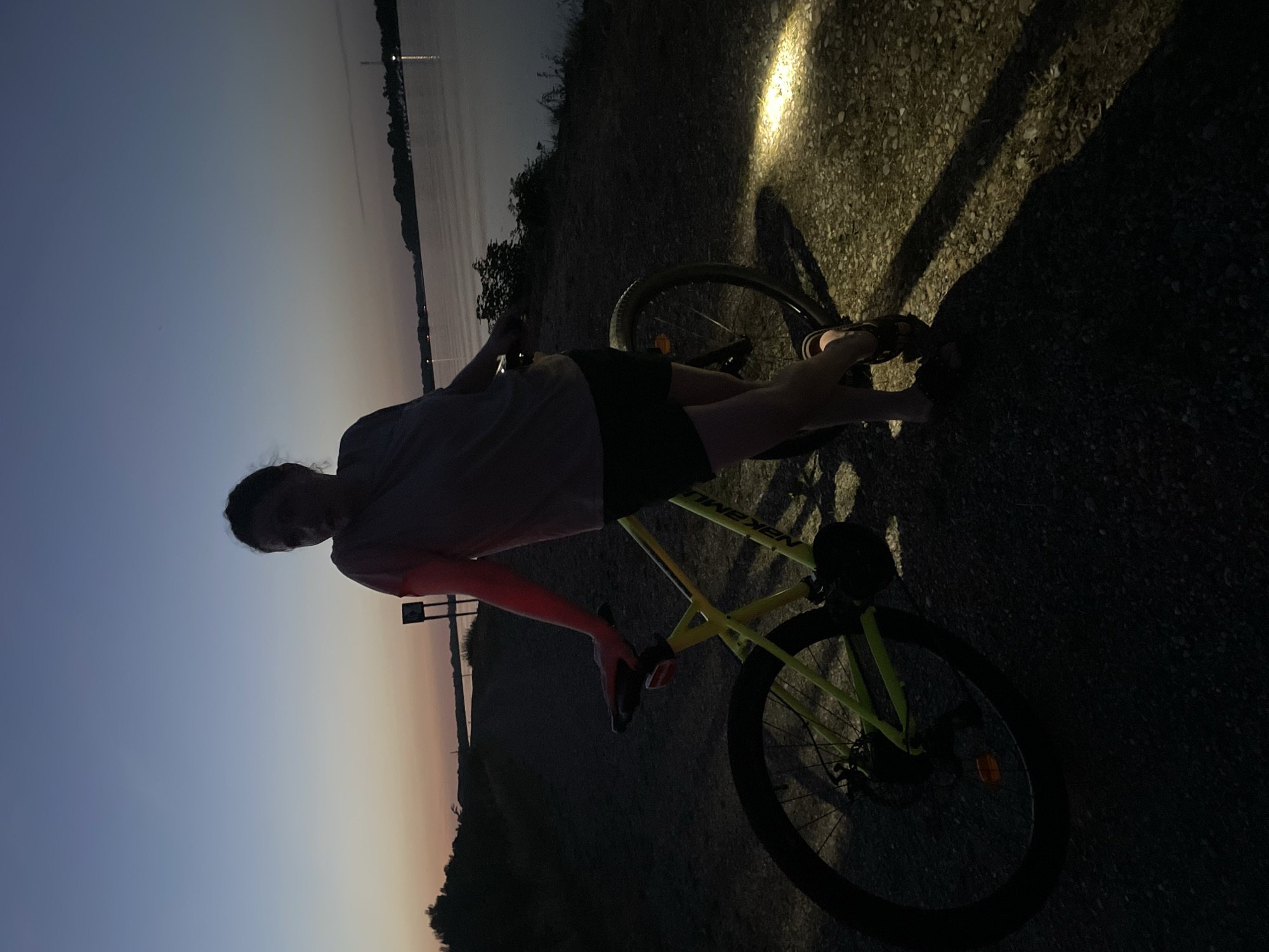 A woman stands with her bicycle near a body of water at dusk. She is wearing a t-shirt and shorts, and is leaning on the handlebars of a bright yellow bicycle. The bicycle has a light on, illuminating the path in front of it. The background shows a body of water, trees, and a darkening sky. The scene appears to be a park or a trail by a river. The woman's posture and facial expression suggest she might be taking a break or posing for a photo. The lighting suggests it is either early morning or late evening. There are no specific landmarks in the image to indicate the precise location within Ottenheim, Germany, but it appears to be a natural area.