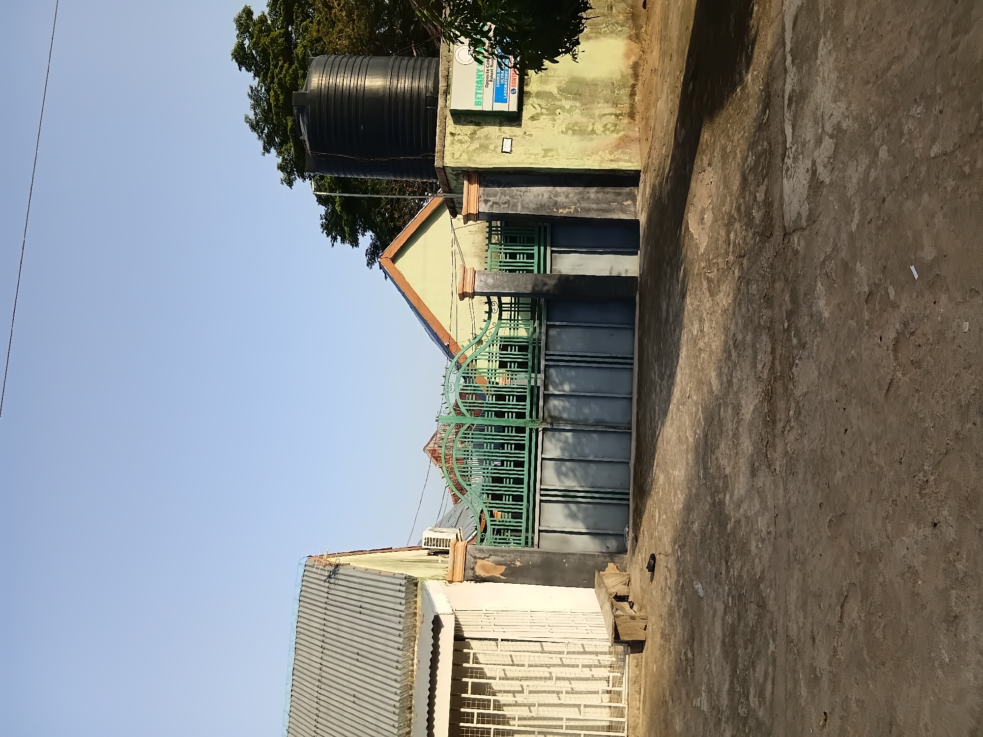The image shows a gate in front of a building in Jalingo, Nigeria. The gate is made of green metal bars and has a gray metal panel underneath. To the left of the gate is a white building with a corrugated metal roof and a white metal security gate on a window. To the right of the gate is a light green building with a large black water tank on top of a concrete structure. A sign with text in blue and white is attached to the concrete structure. The ground in front of the gate is unpaved and appears to be dirt. The sky is clear and blue. The scene appears to be taken during the day, likely in the late afternoon, as indicated by the shadows. There are no people visible in the image.
