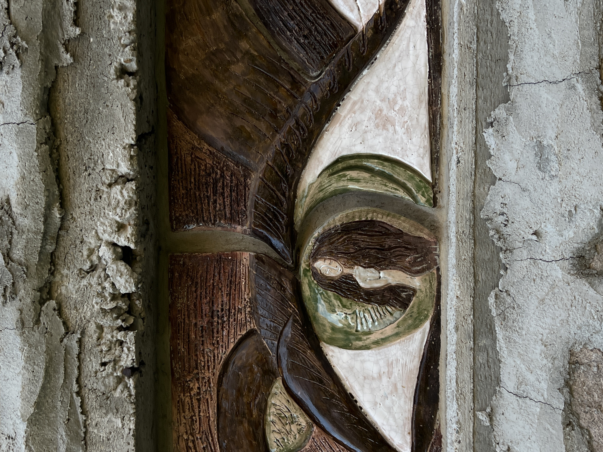 The image shows a close-up of a textured, three-dimensional artistic relief embedded in a rough, concrete-like structure. The relief depicts a large, stylized eye with a green iris. Within the iris, a small, pale-skinned female figure with long, dark hair is visible, seemingly emerging from the eye. The surrounding areas of the relief are textured in shades of brown and white, with detailed lines suggesting eyelashes or feathers. The concrete structure above and below the relief appears weathered, with visible cracks and uneven surfaces. The background is not visible. This artwork is located on the exterior wall of a building in the city of Sinemorets, Bulgaria.