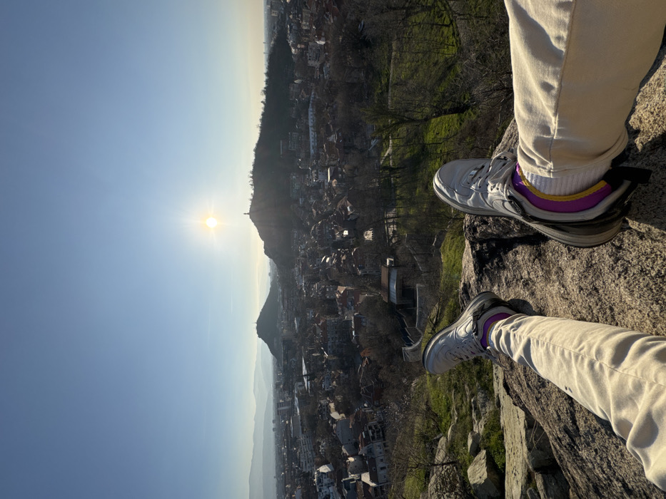 The image depicts a first-person perspective from a rocky vantage point overlooking the city of Plovdiv, Bulgaria, on a clear day with a low sun.
In the immediate foreground, the legs and feet of an individual are extended forward, resting on a large, textured gray rock. The person wears tan-colored trousers, white athletic socks with distinctive purple and yellow horizontal stripes, and light gray athletic sneakers with dark soles. Patches of green grass and sparse, leafless shrubbery are visible on the rocky terrain surrounding the viewer's position.
The midground reveals a panoramic view of the city of Plovdiv. A dense urban sprawl of buildings, many featuring red-tiled roofs, extends across the landscape. Numerous trees, largely bare of leaves, are interspersed throughout the city and on the lower slopes of the hills. A winding path or road with a visible retaining wall is discernable on the hillside directly below the foreground rocks.
In the background, two prominent hills rise above the city. The hill on the right is Bunardzhik Hill, identifiable by the tall, dark silhouette of the Alyosha Monument positioned at its summit. The sun, a bright, radiating orb, is situated directly behind this monument, casting a strong glare and lens flare, and silhouetting the monument. Another, lower urbanized hill is visible to the left. The sky is a clear, light blue, transitioning to a paler hue near the horizon where a distant, hazy mountain range is faintly visible. The low position of the sun suggests either early morning or late afternoon.