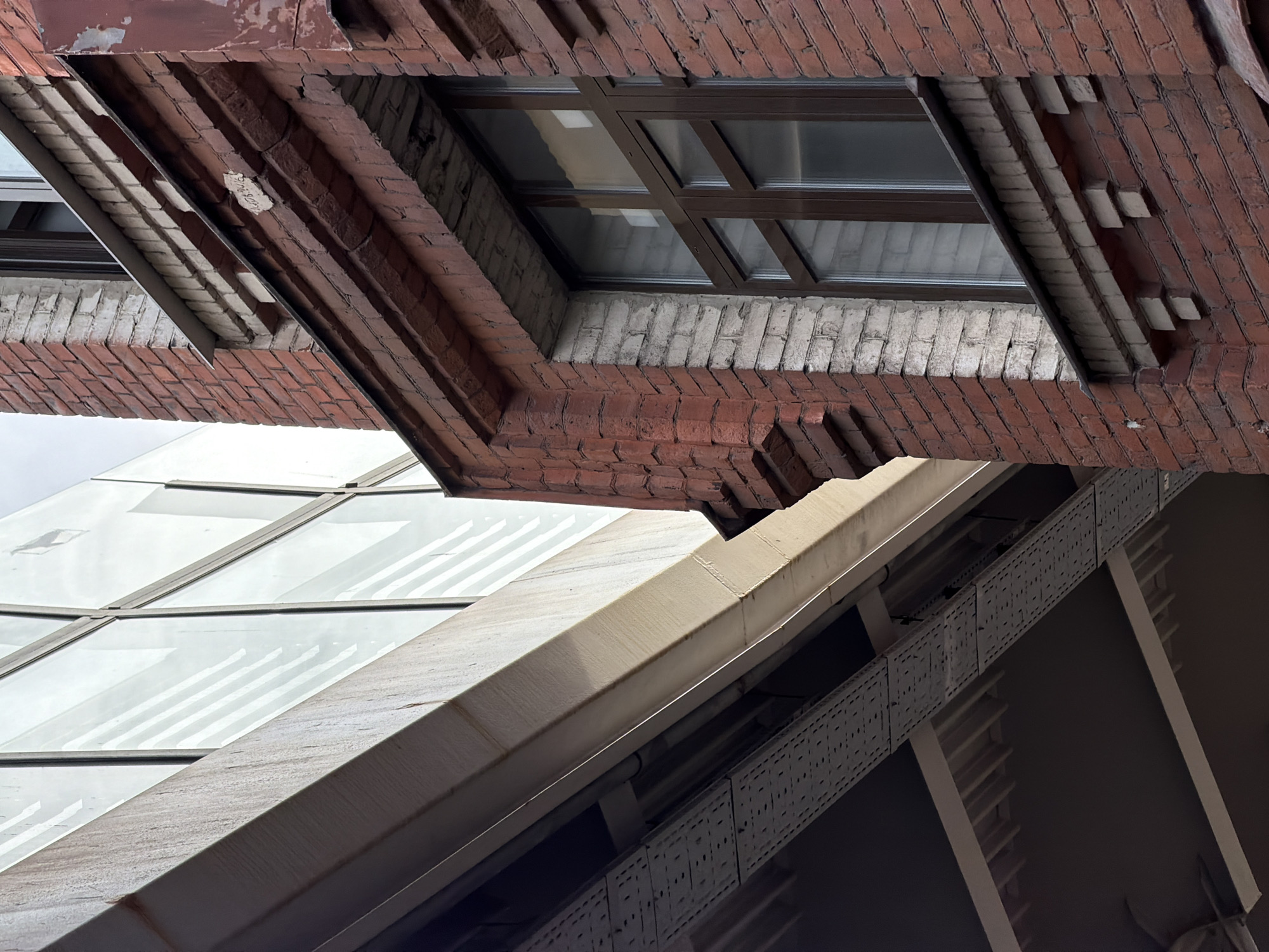 This image, rotated approximately 90 degrees counter-clockwise, presents an upward-looking architectural study in Saint Petersburg, Russia, showcasing a striking contrast between old and new construction. The upper portion of the frame is dominated by a weathered red brick facade, likely part of an older building. An alcove within this brickwork reveals lighter-colored, possibly white or cream-colored, brick and a brown-framed window with multiple glass panes. The window reflects the overcast sky, with some reflections showing subtle patterns. The underside of the red brick overhangs displays some peeling paint or plaster.
Below the older brick structure, a diagonal light-colored architectural element, possibly a concrete beam or stone ledge, crosses the mid-ground. Adjacent to this and extending into the lower right, is a more contemporary structure featuring large glass panels with light-colored frames. These panels reflect linear patterns, suggesting windows or another building nearby. Beneath the glass, a dark, industrial-style metal framework is visible, including sections of perforated metal panels that could be cable trays or part of a walkway.
The lighting suggests an overcast daytime. There are no people visible, and no specific activity is taking place; the image focuses purely on the architectural forms, textures, and the interplay of different construction eras. A faint, illegible white marking, possibly a date or code (e.g., "14.07.2"), is present on one of the red bricks in the upper section of the image.