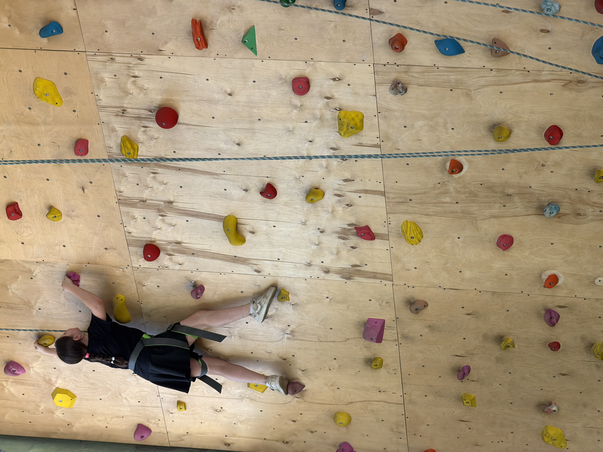 A child wearing a black t-shirt, black shorts, a climbing harness, and white sneakers is rock climbing on a wooden wall with various colored climbing holds. The climbing wall is indoors. A safety rope runs horizontally across the wall. The specific location in Izhevsk, Russia is not discernible from the image.