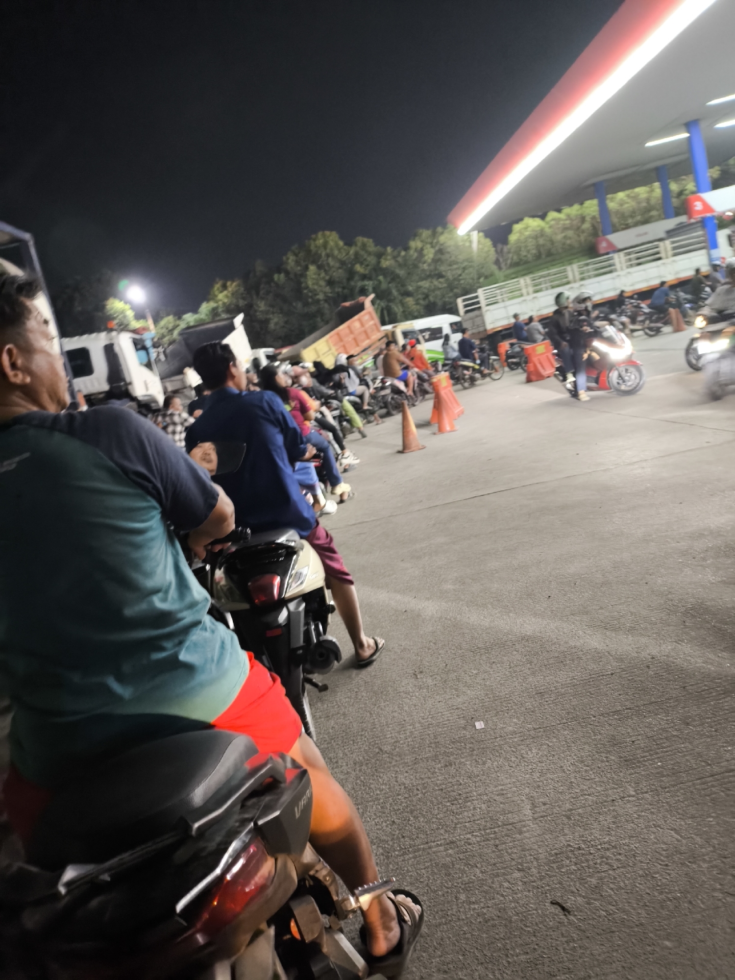 This image depicts a busy night scene at a gas station in Waringinkurung, Indonesia. A long queue of motorcycles and a few trucks are present, indicating a period of high demand or a potential traffic issue. The gas station canopy, illuminated by bright lights, stretches across the upper portion of the frame, with a red and white stripe along its edge. Trees are visible in the background, silhouetted against the night sky. Several individuals are seen on or around their motorcycles, some appearing to be waiting in line. The ground is paved with concrete, and orange traffic cones are placed along the queue. The overall atmosphere suggests a normal, albeit busy, operational night at this location.