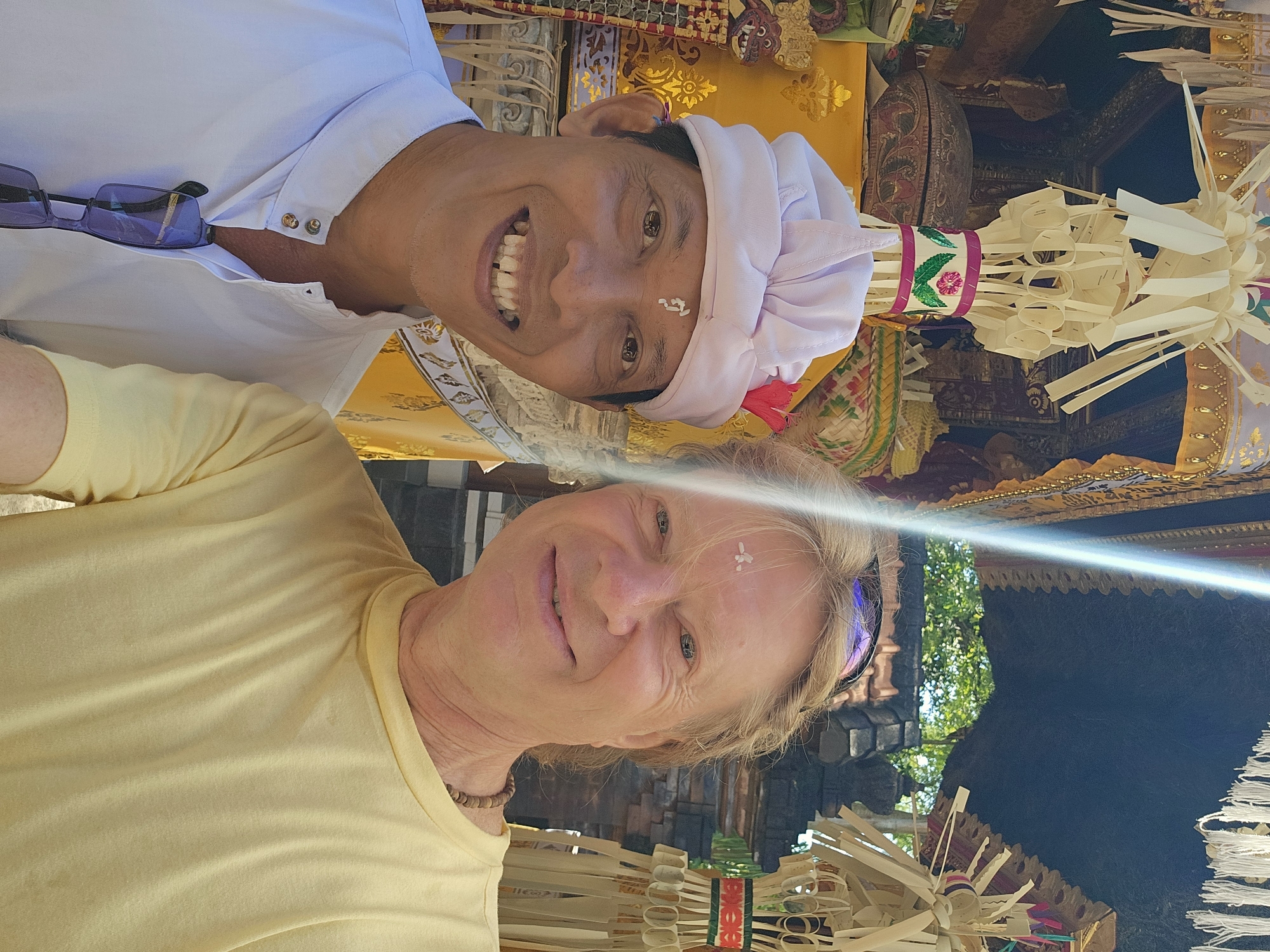 A selfie captures two smiling individuals in what appears to be a Balinese ceremonial or temple setting in Denpasar, Indonesia. The image is oriented vertically, with the subjects looking towards the camera.
In the background, a man with a wide smile is visible, facing the camera. He has dark hair, dark eyes, and a small white mark on his forehead, likely *bindu* (rice paste) from a blessing. He is wearing a white collared shirt and a traditional white Balinese *udeng* (head covering) adorned with a small red flower. A pair of sunglasses is tucked into his shirt collar. Behind him, intricate gold and colorful fabrics, typical of Balinese temple decorations, are visible, along with woven palm leaf offerings.
In the foreground, a woman with blonde hair is smiling gently, also facing the camera. She wears a light yellow long-sleeved top and a simple beaded necklace. Similar to the man, she has a small white mark on her forehead. Sunglasses are pushed up onto her head, reflecting sunlight.
The setting is richly decorated with traditional Balinese elements, including elaborate woven palm leaf ornaments and colorful ribbons (pink, green), suggesting a religious ceremony or cultural event. Glimpses of a dark, ornate structure and some greenery can be seen in the background. The bright lighting indicates it is daytime, likely sunny. Both individuals appear happy and engaged in the moment. There is no visible text in the image.