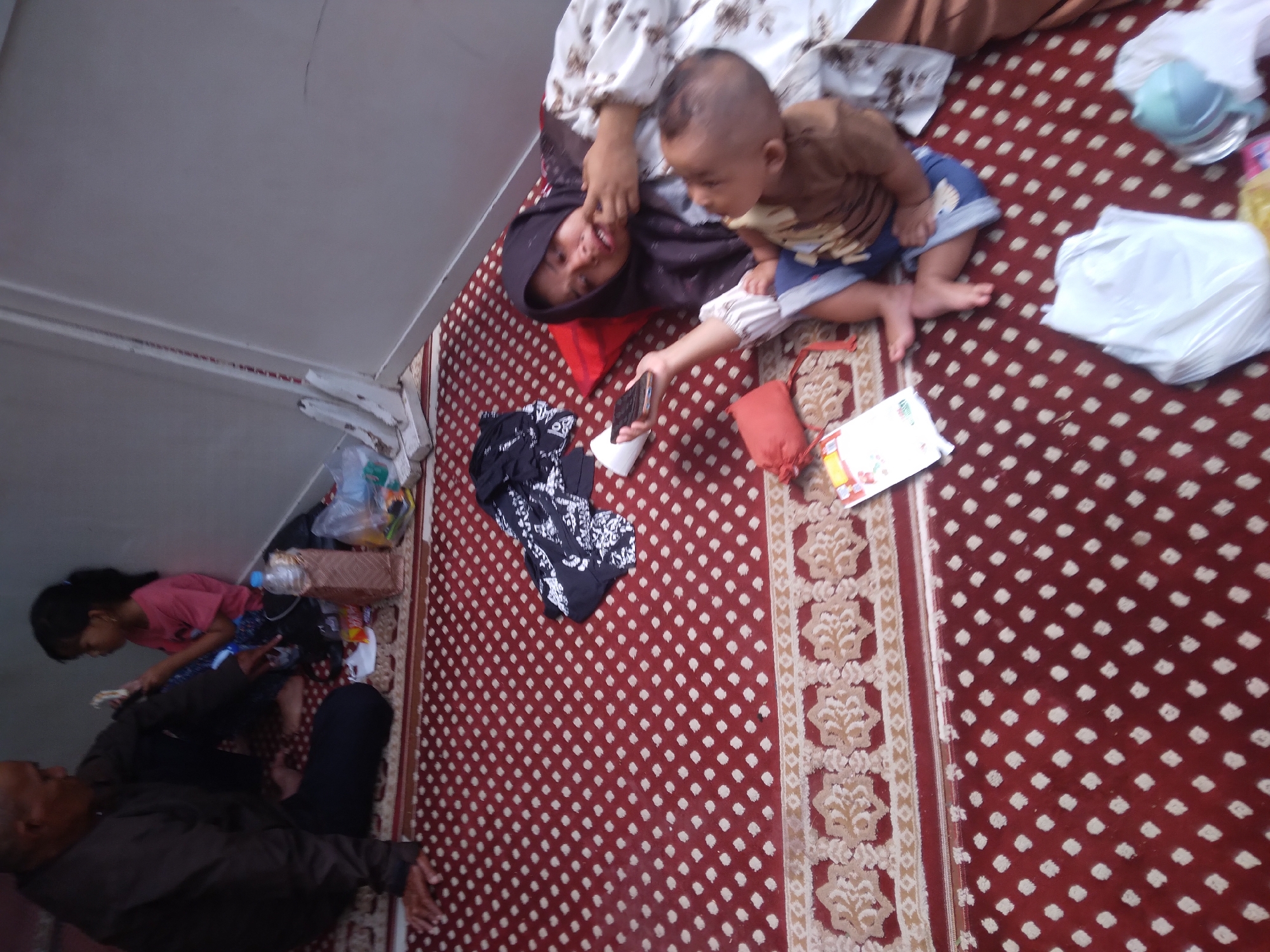 A group of people are sitting on a red carpeted floor. In the foreground, a woman wearing a hijab and a white patterned dress is lying down and holding a smartphone. A young child, wearing a brown shirt and blue shorts, is sitting next to her and looking at her. Further back, an elderly man in a dark jacket is sitting and interacting with a young girl in a pink shirt. Scattered on the carpet are various items including a red bag, a white paper, a bottle of water, and plastic bags. The scene appears to be indoors, possibly in a makeshift shelter, with a white wall in the background. The setting is likely Desa Pamijen, Kecamatan Sokaraja, Indonesia.
