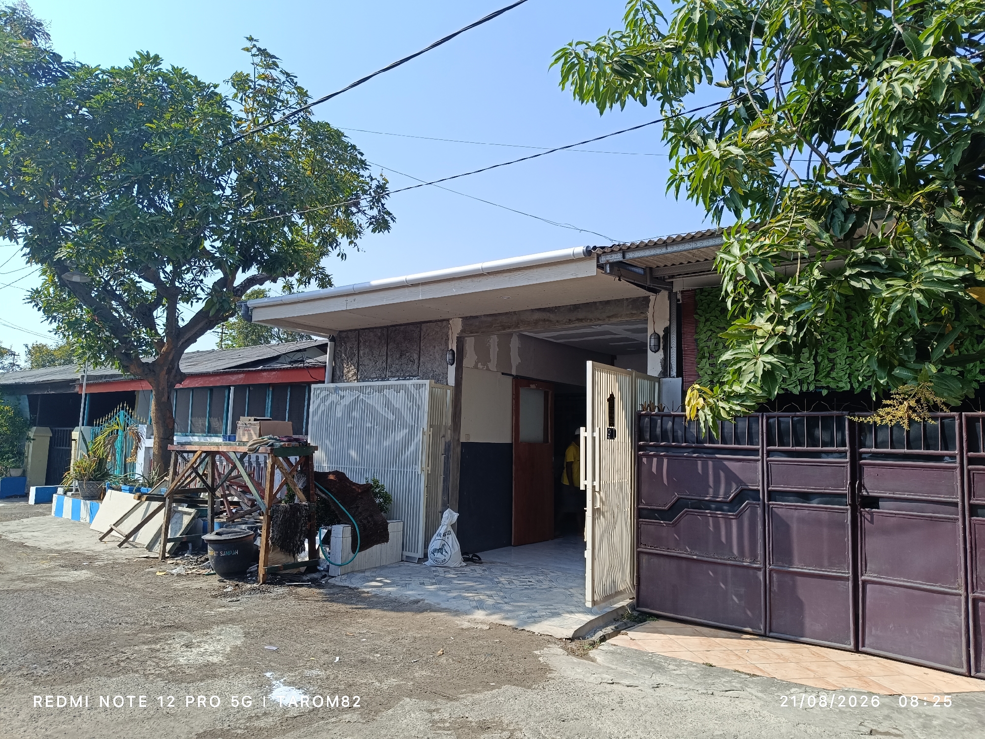In the city of Sruni, Indonesia, this image captures a scene of a residential property with a large tree in the foreground, casting shadows on the street. The property features a brown metal gate and a white gate, both appearing to be closed or partially open. The house itself has a visible roof structure and walls constructed from a combination of stone and what appears to be concrete. In front of the house, there's an open entrance leading into a covered area, with a person in a yellow shirt visible inside. Adjacent to the entrance, a white bag with text is placed on the tiled ground. The surrounding environment shows a paved street with some debris, and several smaller plants and potted plants are scattered around the property. A wooden structure with various items on it, including what looks like a trash can with text, is situated to the left of the entrance. Power lines are visible crisscrossing the clear blue sky above. The image was taken on August 21, 2026, at 08:25.