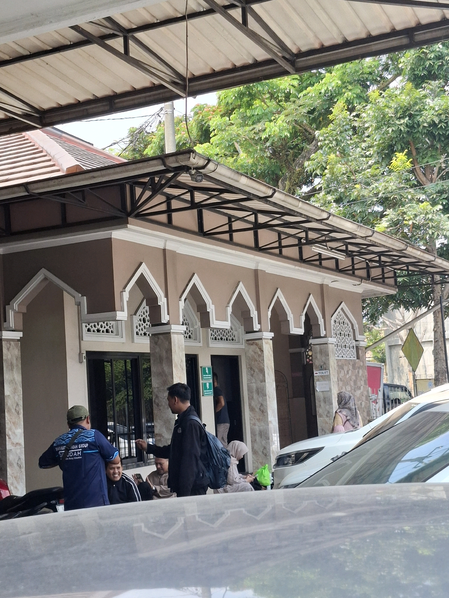 The image depicts the exterior of a building, likely a public restroom facility, with an architectural style featuring arched entrances and decorative latticework. The building's facade is painted in a light brown color, with beige and grey stone accents. Several people are visible in the foreground and near the entrance. One man in a blue shirt with the text "SUJUD" is standing and gesturing. Another man in a black jacket is facing him, holding a backpack. Two individuals in the background appear to be sitting or standing near the entrance. A white car is parked in front of the building. The scene is set outdoors, with trees and electrical wires visible in the background. A sign with the word "TOILET" is mounted on the wall. The lighting suggests it is daytime, with the sun casting shadows. The image's context, including the sign and the general appearance of the building, suggests it could be a rest area or facility located in Sukabumi, Indonesia, potentially along a travel route or in a public space.
