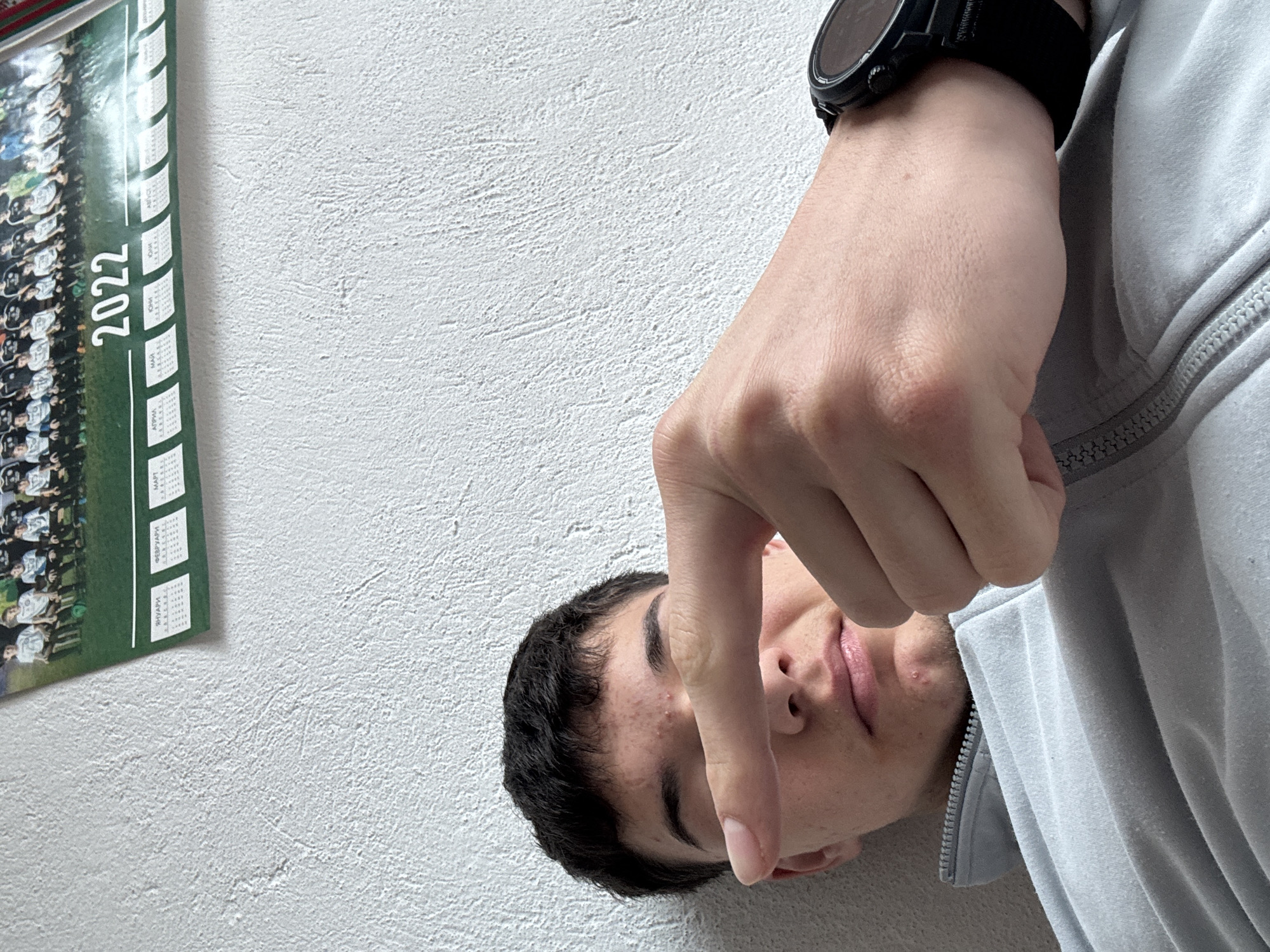 A man wearing a black wristwatch is holding his index finger over his face. The image includes a calendar that reads "2022" in Cyrillic script. The location is unknown and cannot be determined.