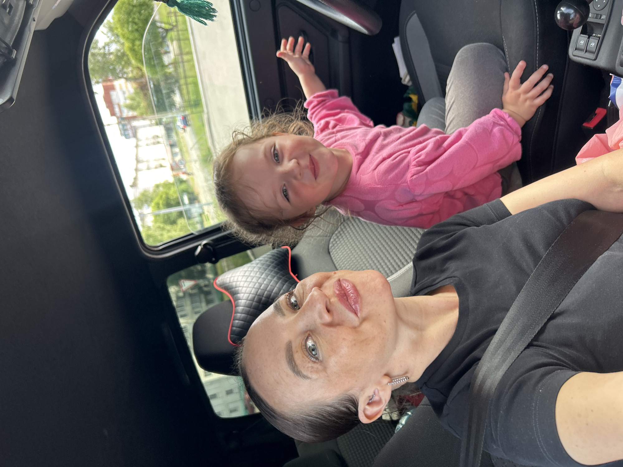 In the image, a woman and a young girl are sitting in the front seats of a car. The woman is in the foreground, taking a selfie. She has dark hair slicked back, light-colored eyes, and is wearing a black t-shirt and a seatbelt. The girl is in the background, wearing a bright pink, long-sleeved sweater with polka dots. She has light brown curly hair and blue eyes, and appears to be smiling and looking at the camera with her hand raised. The car has a dark interior, with a sunroof visible above the passengers. Through the windows, greenery and buildings are visible, suggesting an urban or suburban setting. The weather appears to be overcast or cloudy, as the light is diffused. The overall mood of the image seems happy and casual, with the two subjects appearing relaxed and engaged with each other. The photo was taken in Chaikovsky, Russia. There is no visible text in the image.