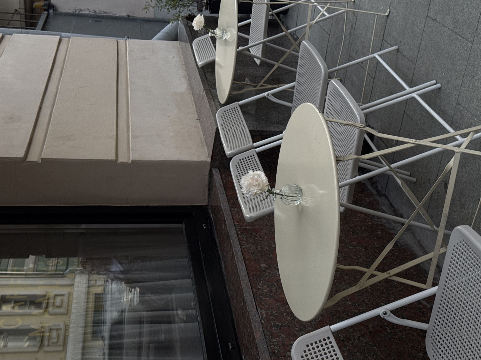This image depicts an empty outdoor cafe setting, viewed from an elevated perspective looking downwards. The scene is rotated 90 degrees clockwise.
In the mid-ground, a series of light-colored (off-white or beige) oval tables are arranged, each accompanied by matching chairs. The chairs have perforated seats and backs, typical of contemporary outdoor furniture. Each table features a small, clear glass vase holding a single white flower, possibly a carnation, adding a delicate touch to the unoccupied space. The tables and chairs appear to be made of metal, suggesting durability for outdoor use.
To the left, a portion of a building's facade is visible, constructed from a light-colored material with recessed vertical lines. Below this, a large, dark-framed window reflects parts of buildings opposite, including windows and architectural details, although no specific text is legible in the reflection.
The ground beneath the cafe setup is paved. Closer to the building, it appears to be a reddish-brown speckled stone, possibly granite, transitioning to a darker gray, possibly cobblestone or block paving, further into the background.
The lighting is soft and diffused, characteristic of an overcast day, as there are no harsh shadows. The absence of people creates a quiet and serene atmosphere. No discernible text is visible on any of the objects or surfaces. The setting suggests an urban environment, specifically an outdoor cafe in Moscow, Russia.