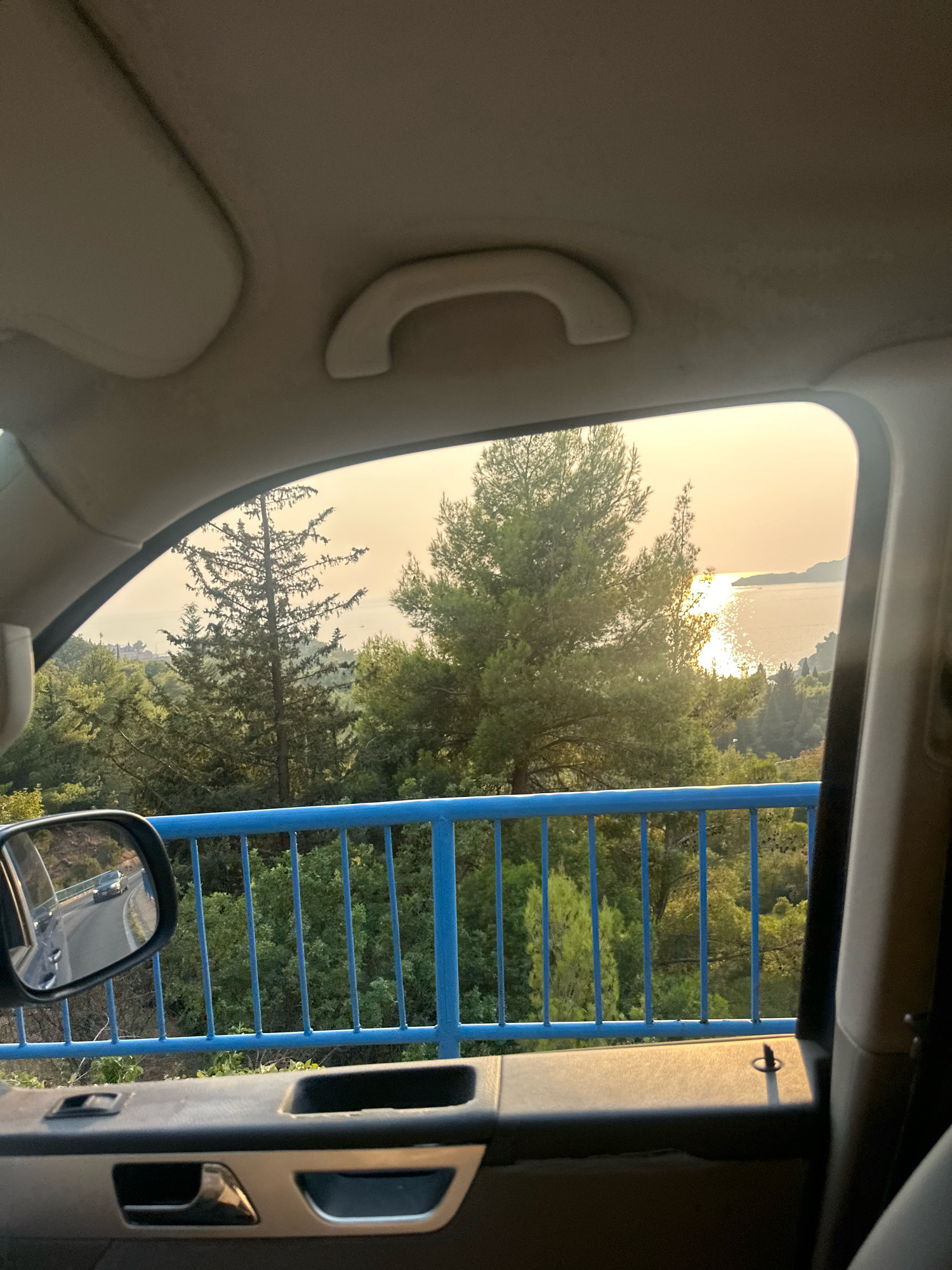 The image shows a view from inside a vehicle looking out a window. The foreground features the interior of the car, including a door handle, window controls, and a portion of the ceiling with a grab handle. A blue railing runs horizontally across the lower part of the view outside the window. Beyond the railing, a dense green forest with various trees stretches towards a body of water, likely the sea, in the distance. The sun is low in the sky, casting a warm, golden light and reflecting off the water, suggesting either sunrise or sunset. A side-view mirror attached to the car door reflects a winding road with another vehicle. The location is identified as Podličak, Montenegro.