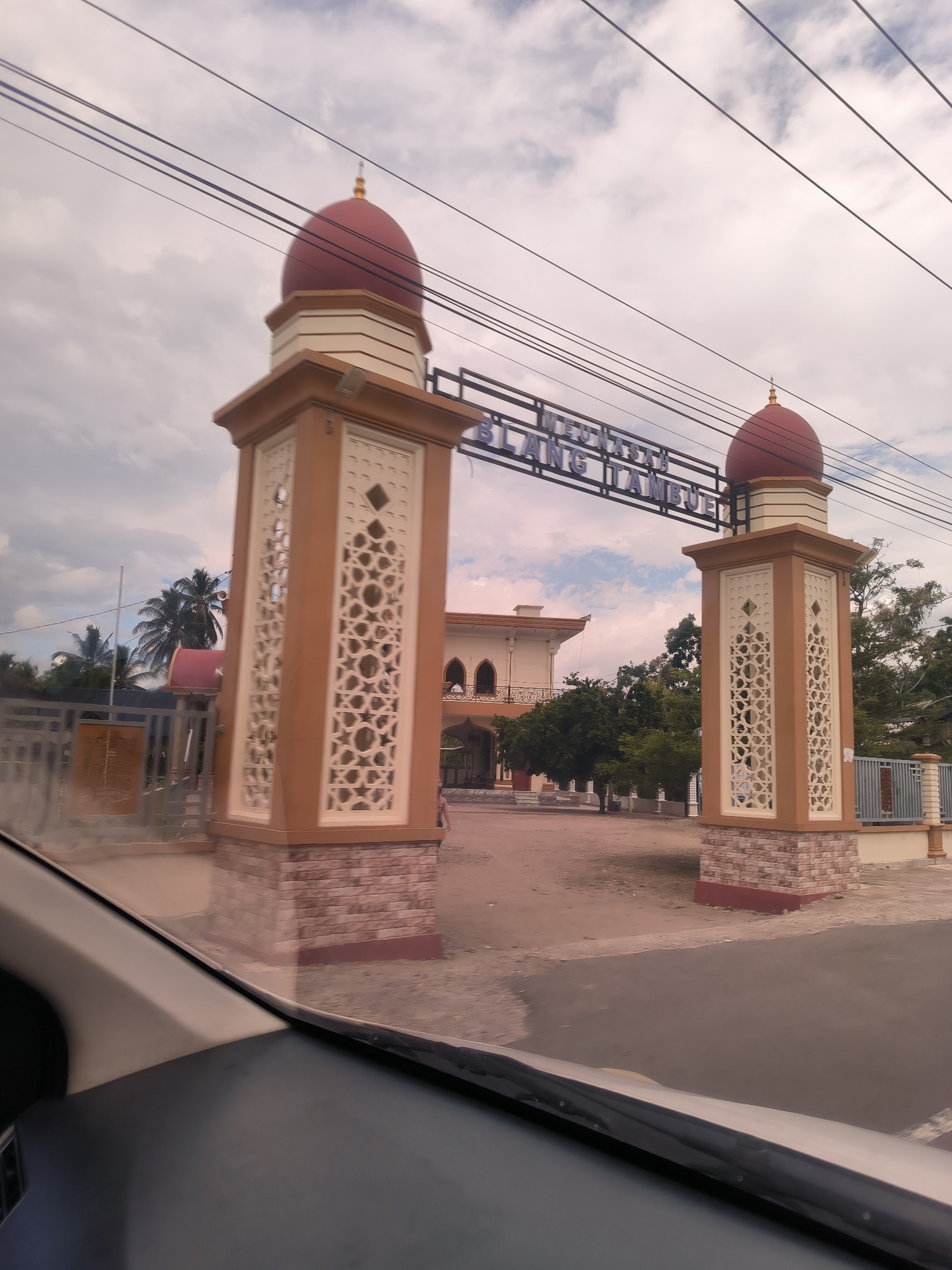 The image, taken from inside a vehicle looking out the windshield, depicts an ornate entrance gate under a cloudy sky. The bottom of the frame shows the car's dashboard and a reflection on the windshield.
In the midground, a large gate stands prominently, composed of two substantial, light-brown pillars. Each pillar is adorned with intricate, cream-colored geometric lattice work and features a reddish-brown brick-like base. Topping each pillar is a distinct red, domed cupola finished with a golden finial. A horizontal sign stretches between the two pillars, displaying blue lettering that reads "MEUNASAH BLANG TAMBUE." Overhead, several power lines run across the sky, partially obscuring the sign and the clouds.
Beyond the gate, a multi-story building with a light-colored facade, arched windows, and balconies is visible, surrounded by lush green trees and some palm trees on the left. The architectural style, particularly the domes and geometric patterns, suggests an Islamic religious or community structure.
The scene appears to be set during the daytime, with an overcast sky indicated by the soft, diffused lighting and visible clouds. No individuals are clearly discernible, and the primary activity captured is observing the entrance from a moving or stationary vehicle. The location cues, especially the visible text, place the scene in Blangtambue, Indonesia, likely at a "Meunasah" (a small mosque or prayer house).