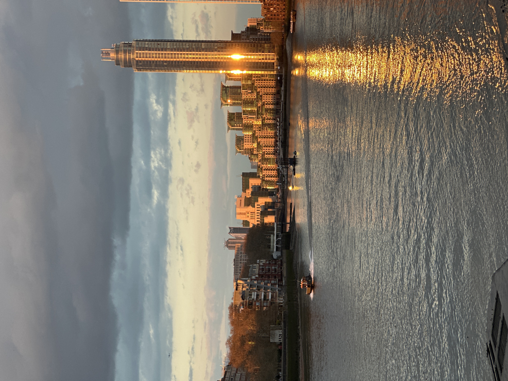 The image depicts a cityscape in London, United Kingdom, at sunset, with a river running vertically through the center. On the right, a tall skyscraper reflects the setting sun, which creates a bright, golden glare on the river's surface. Other buildings of varying heights line the riverbank. A boat moves along the river, visible in the lower part of the frame. The left side of the image shows a cloudy sky, transitioning from dark grays to lighter hues near the horizon.