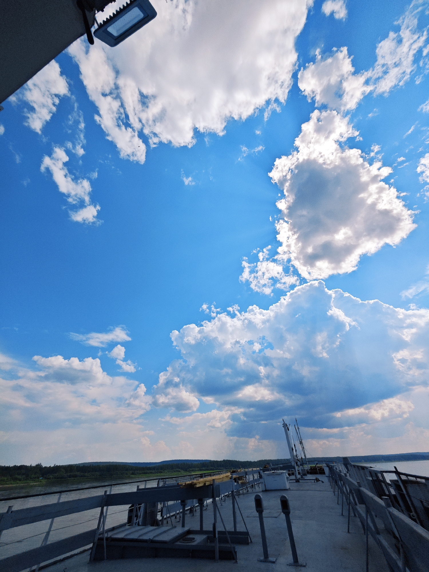 This media file captures a low-angle shot of a bright blue sky filled with fluffy white clouds, some of which are lit by sunbeams breaking through. In the foreground, the deck of a vessel is visible, with metal railings and various equipment such as poles and what appears to be a white utility box. The deck is constructed of concrete. In the distance, a tree-lined shore stretches out along a body of water, with rolling hills forming the horizon under the expansive sky.
The scene suggests a daytime setting, likely late morning or afternoon, given the bright sunlight and clear sky. The weather appears fair with some scattered clouds. The location cues, particularly the river and the dense forest on the distant banks, combined with the provided context of Podkamennaya Tunguska, Russia, point to a natural, possibly remote environment. There are no people visible in the media. The overall mood is serene and expansive, emphasizing the vastness of the sky and the natural landscape. There is no visible text in the media file.