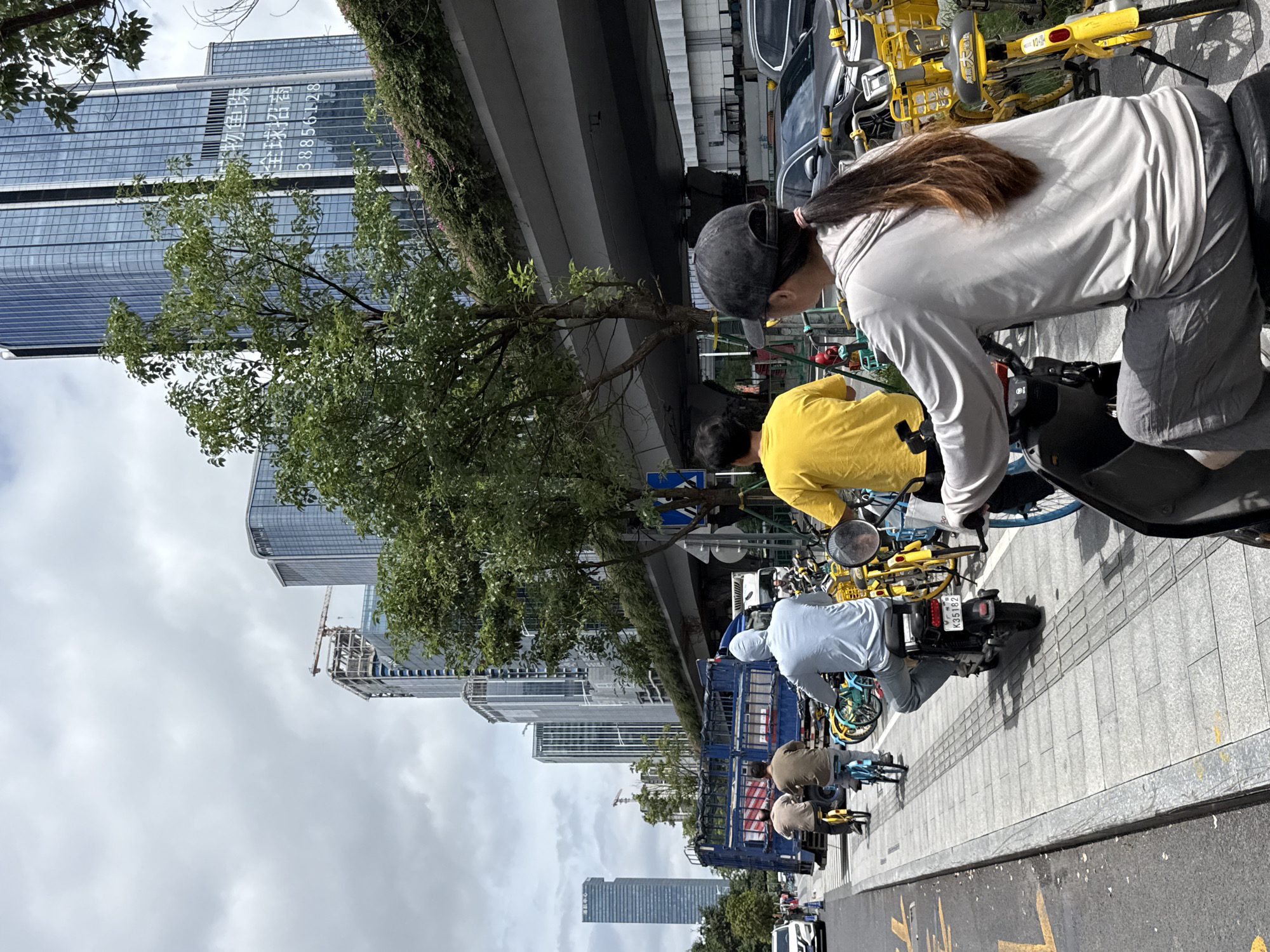 In Tianhe District, China, under an overcast sky, a street scene unfolds with multiple people riding motorcycles and bicycles. In the foreground, a person wearing a grey hoodie and grey pants rides a scooter. Behind them, a person in a bright yellow shirt is also on a scooter. Further down the street, three individuals on motorcycles are riding on a paved sidewalk. A blue truck is parked on the left side of the road. Numerous yellow and blue rental bicycles are parked along the right side of the sidewalk. Tall modern buildings dominate the background, with a highway overpass visible above the street. The environment is urban, with signs of construction and development.