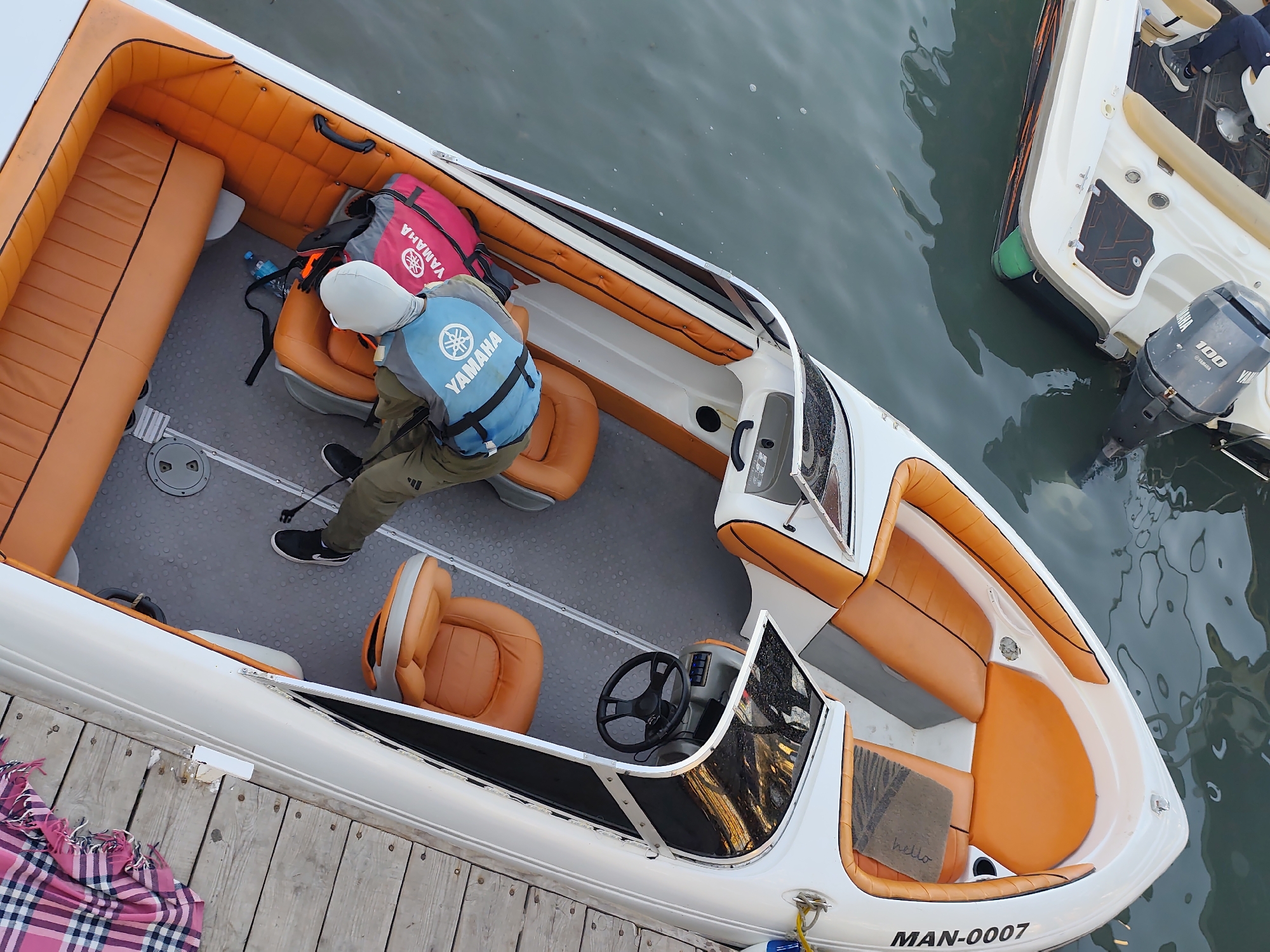 This is a high-angle, eye-level shot of a white speedboat with bright orange upholstered seating. The boat is docked next to a wooden pier, where a pink and blue plaid blanket is draped over the edge.
Inside the speedboat, a person wearing a light gray hoodie, olive green pants, black athletic shoes, and a blue Yamaha life vest is seated. They are wearing a white hooded mask that covers their face. A pink Yamaha life vest is placed behind them on the seat, along with a dark backpack. There's also a blue plastic water bottle on the floor. The boat has a steering wheel and console area, and more orange seating is visible in the front. Another white speedboat with orange accents is partially visible in the background, also in the water.
The scene appears to be taking place outdoors, with the water reflecting the overcast sky, suggesting a cloudy day. The calm water indicates no significant waves or strong currents. The overall mood is relaxed and possibly preparing for or just finished a boat trip. Visible text includes "YAMAHA" on the life vest and on the pink vest, "100" on the outboard motor, and "MAN-0007" on the stern of the boat. There is also a small welcome mat with the word "hello" on it.