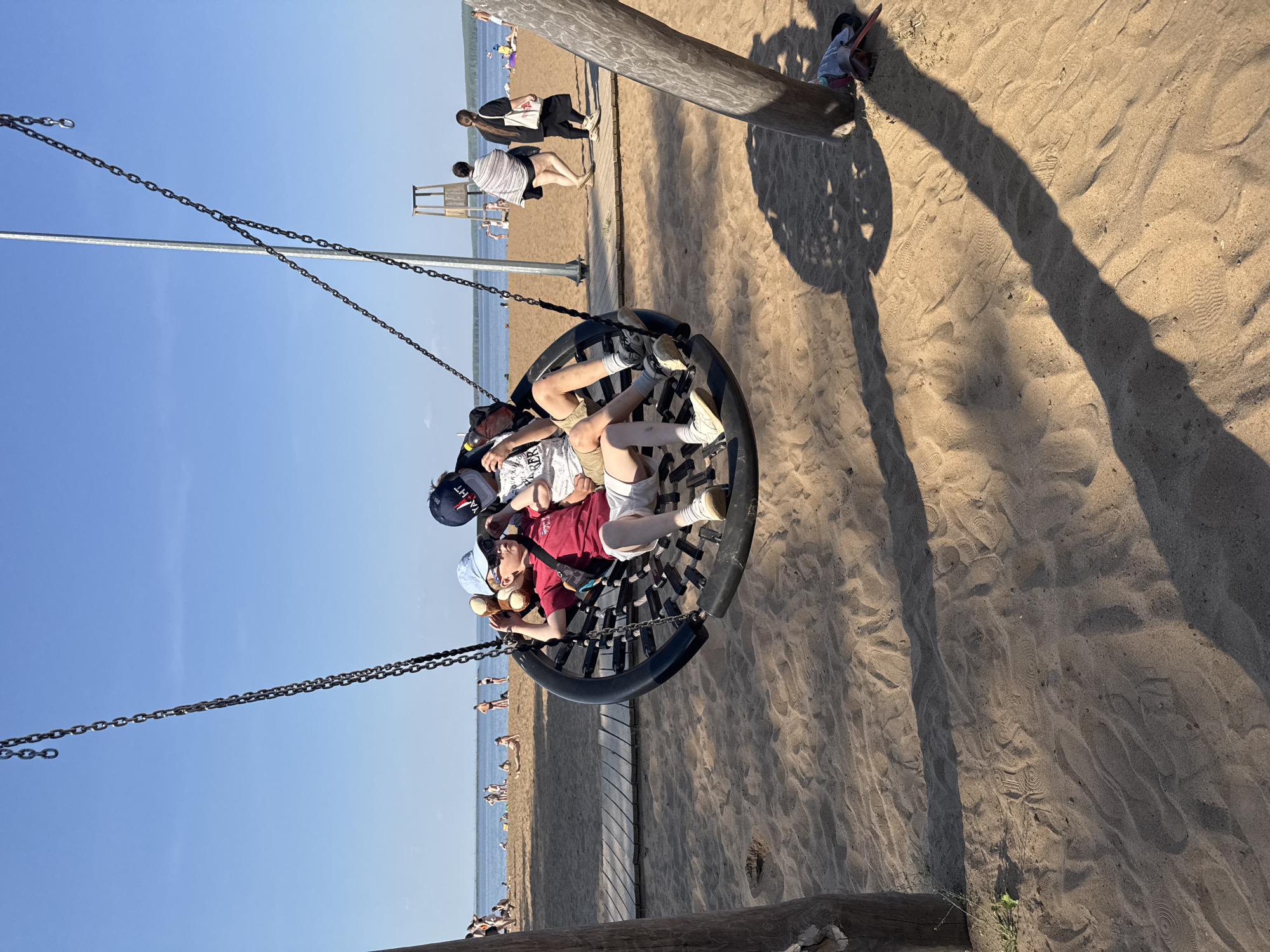 Three children are on a circular swing at a beach in Chaikovsky, Russia. The swing is suspended by chains from a wooden beam. Two of the children are lying down in the swing, and one is sitting up. The child who is sitting up is holding a teddy bear. The beach has sand and a wooden walkway. People are walking in the background. The sky is blue.