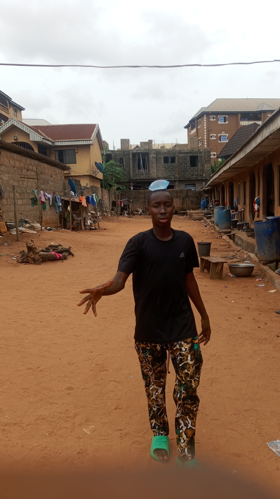 The image depicts a young man in the city of Nkpor, Nigeria. He is standing in a sandy, unpaved area with buildings on either side. The man is wearing a black t-shirt, patterned trousers, and green sandals. He has a blue object on his head. His right arm is extended with an open hand. In the background, there are buildings of varying construction quality, some with satellite dishes. Laundry is hanging on a line near one building. There are several large blue barrels, a metal basin, and a wooden bench along the side of one building. A pile of wood and debris is visible in the background. The sky is overcast.