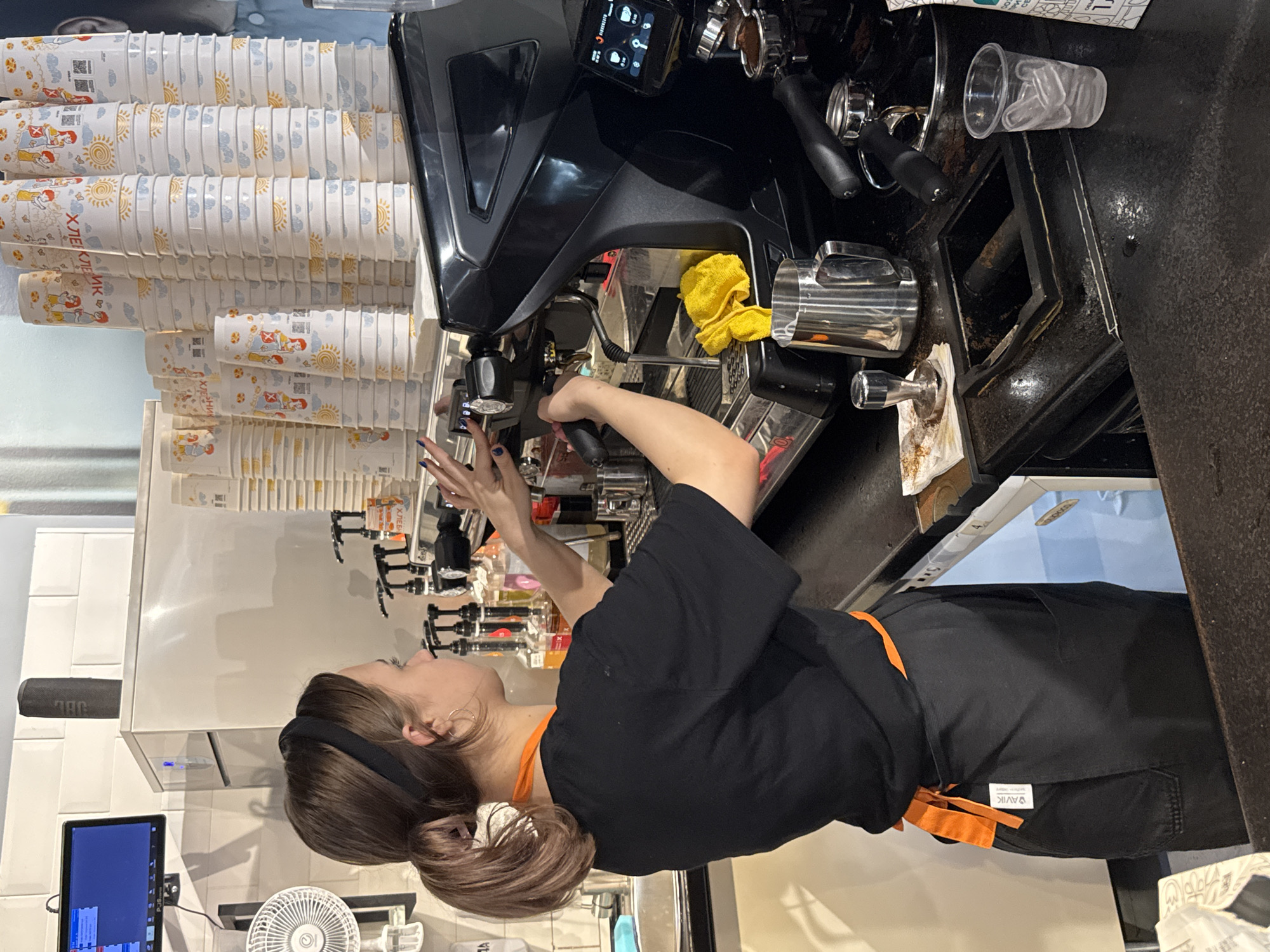 An image taken in a coffee shop in Saint Petersburg, Russia, shows a female barista with brown hair tied back in a ponytail, wearing a black t-shirt and an orange apron. She is standing behind a black commercial espresso machine, in the process of preparing a beverage. Her hands are near the machine's controls and portafilter. Several stacks of disposable paper cups are visible in the background, along with bottles of syrups. A stainless steel milk jug and a tamper are on the counter in the foreground, along with a clear plastic cup filled with ice. A small fan is mounted on the wall to the left. A monitor displaying what appears to be a point-of-sale system is also mounted on the wall. The environment suggests a professional food service setting.