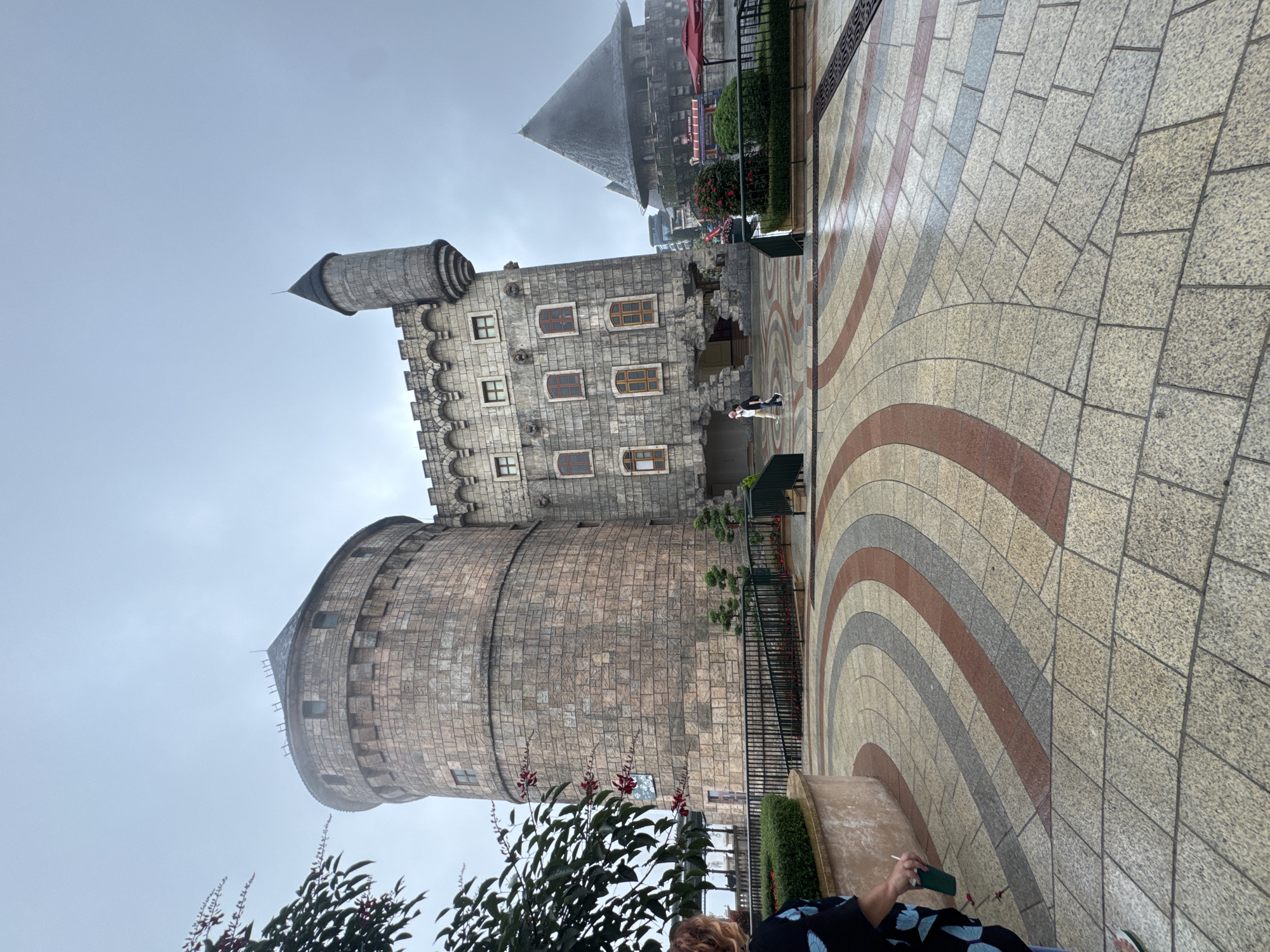 The image depicts a castle-like structure in Đà Nẵng, Vietnam. The main building is constructed of light-colored stone, with a large cylindrical tower on the left, topped with a conical roof. Adjacent to the tower is a rectangular structure with multiple window openings. In the background, a taller tower with a pointed roof is visible. The foreground features a paved area with a curved pattern of light and dark stones. Part of a person is visible in the lower right corner. The sky is overcast and appears foggy. Vegetation including bushes and small trees can be seen in the foreground and around the base of the castle.