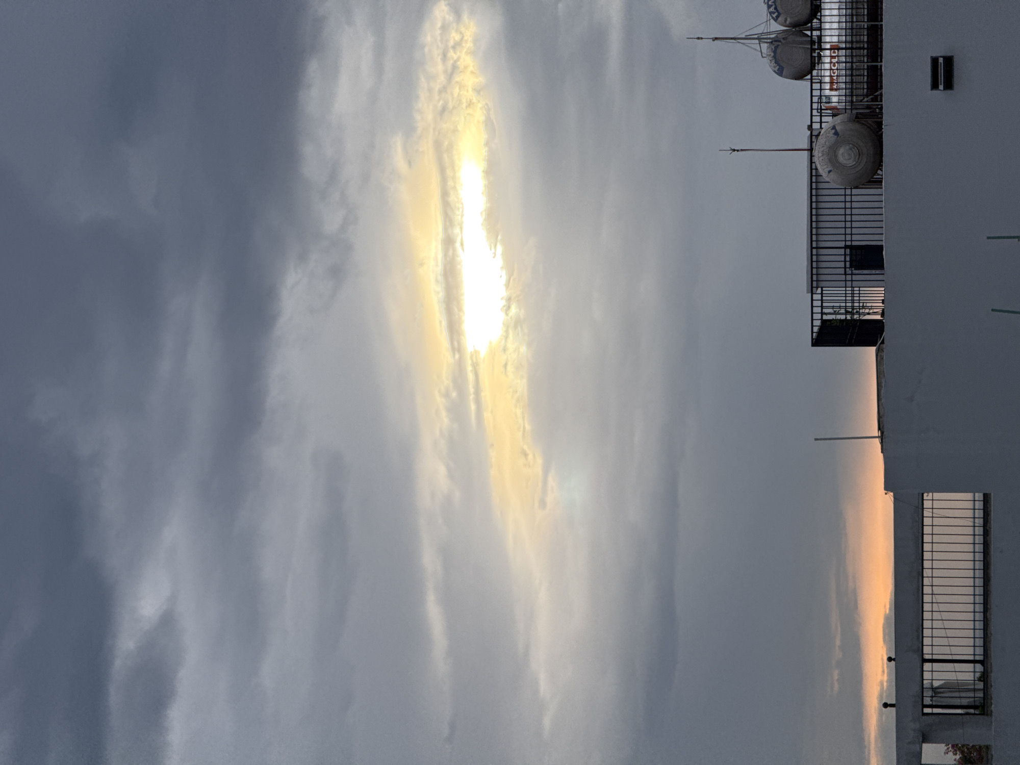 A cloudy sky at dusk is visible over a rooftop in Đà Nẵng, Vietnam. A sliver of sunlight breaks through the dense cloud cover, creating a bright, illuminated band across the sky. Below, the rooftop is equipped with water tanks and a security fence. A small window is visible on the side of a building. The lower portion of the image shows a railing and part of another building. The sky transitions to a soft orange glow near the horizon.