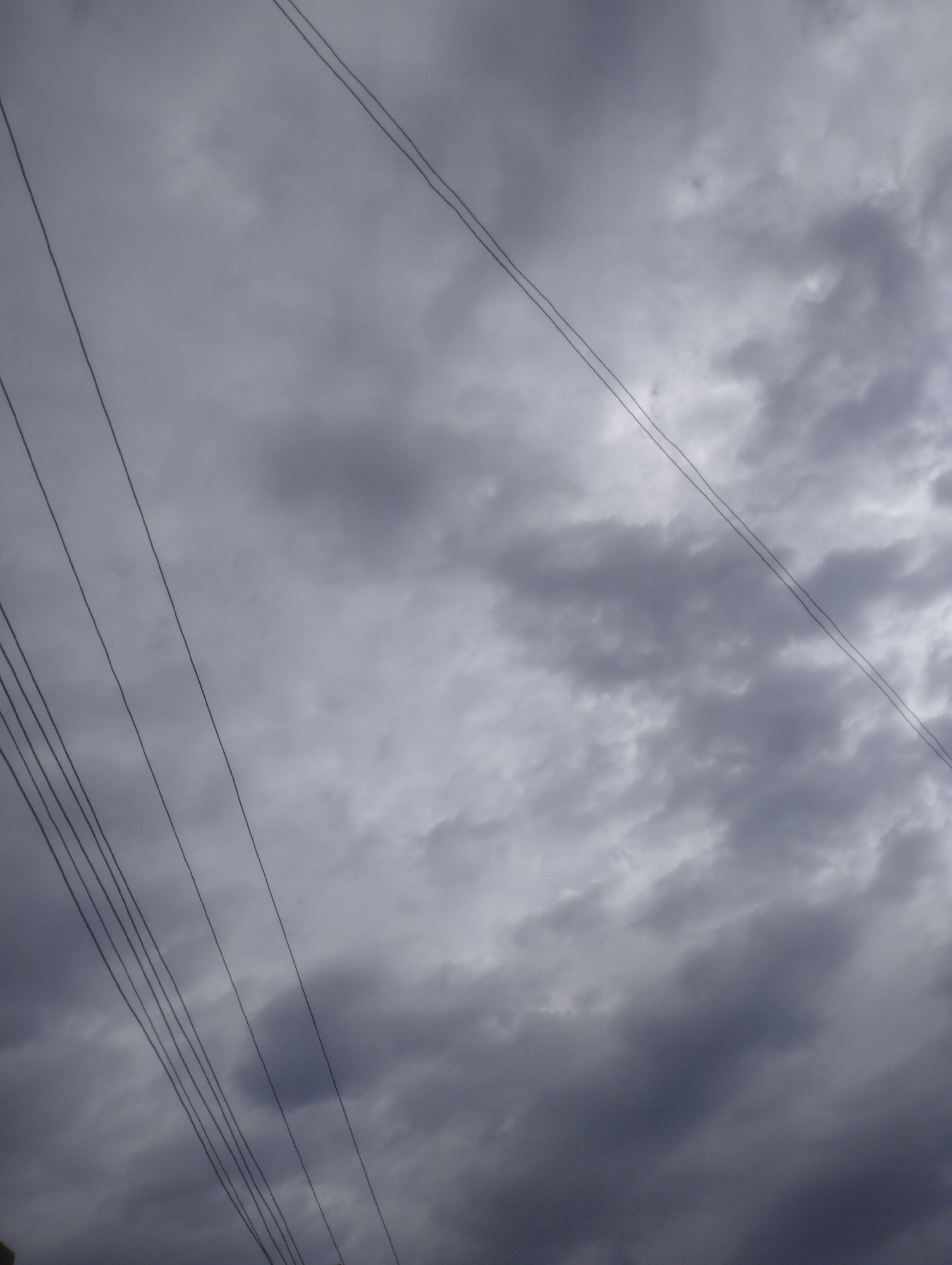 The image captures a cloudy sky with multiple power lines stretching across the frame. The clouds are varied in shade, suggesting an overcast or possibly stormy condition. The power lines are dark and appear to be suspended high above, silhouetted against the lighter sky. The scene is devoid of any people or specific landmarks, making it impossible to determine a precise location within Jos, Nigeria, or any other city. The focus is solely on the atmospheric conditions and the man-made infrastructure of the power lines.