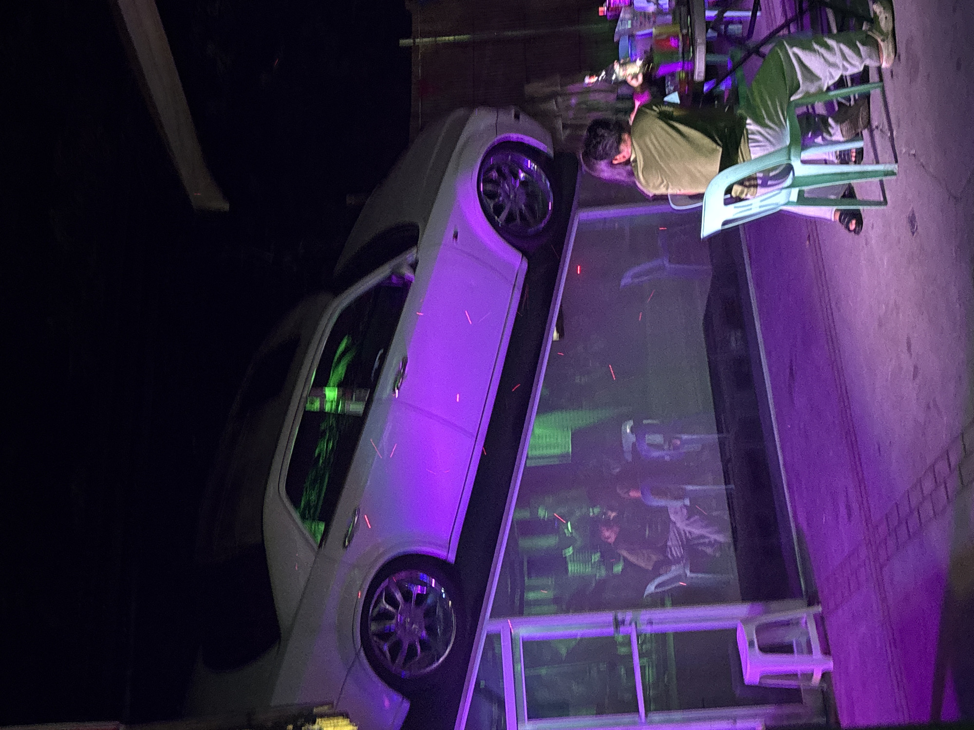 A white car is mounted on a structure above a glass-enclosed area. The car is illuminated with purple and green lights. Below, in the glass-enclosed area, several people are visible sitting at tables. Outside the glass enclosure, one person sits in a mint green chair, facing away from the camera, towards a table with drinks. The ground is paved with bricks and illuminated with purple light. This scene appears to be an outdoor establishment or street-side venue in Davao City, Philippines, characterized by its unique car display and vibrant lighting.
