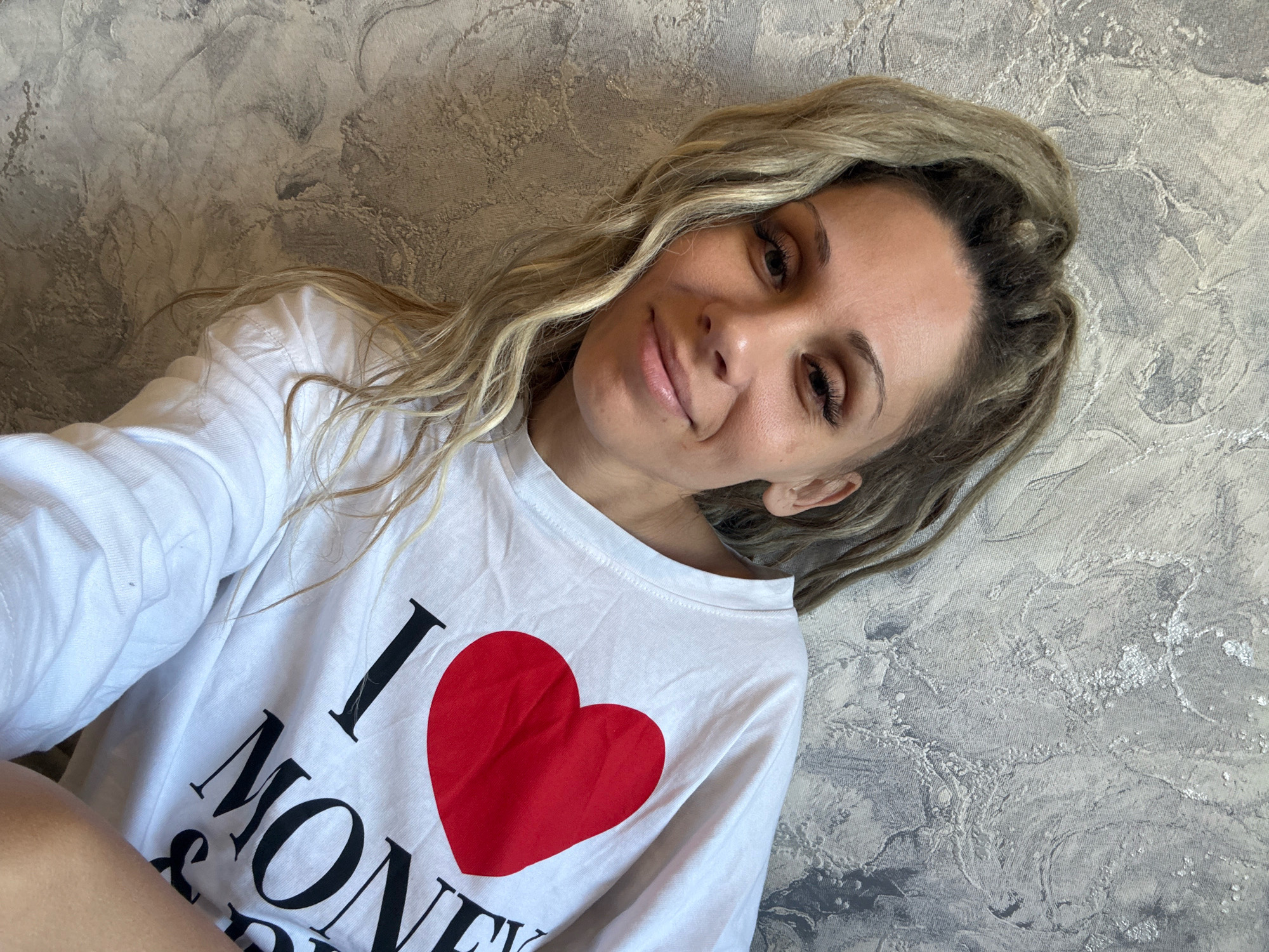 A woman with blonde hair is taking a selfie while leaning against a textured gray wall. She is wearing a white long-sleeved shirt with a large red heart and black text that reads "I LOVE MONEY". The woman is smiling and looking at the camera. The background wall has a marbled or textured pattern. The image is cropped to focus on the woman and her shirt.