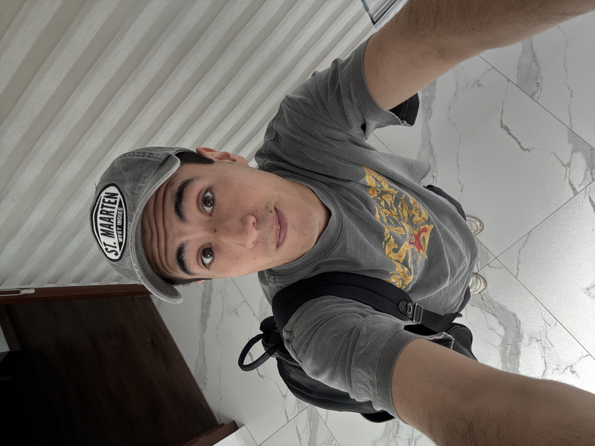 The image is a high-angle, selfie-style photograph of a young man wearing a grey t-shirt and a grey baseball cap with "ST. MAARTEN WEST INDIES" embroidered on it. He is also wearing a black backpack. He is standing on a tiled floor with a marble-like pattern. To the left of the frame is a dark wooden surface. In the background, there is a wall with horizontal blinds. The man is looking directly at the camera with a slightly surprised expression. The lighting suggests an indoor environment. The location is identified as San Bernardino Tlaxcalancingo, Mexico.