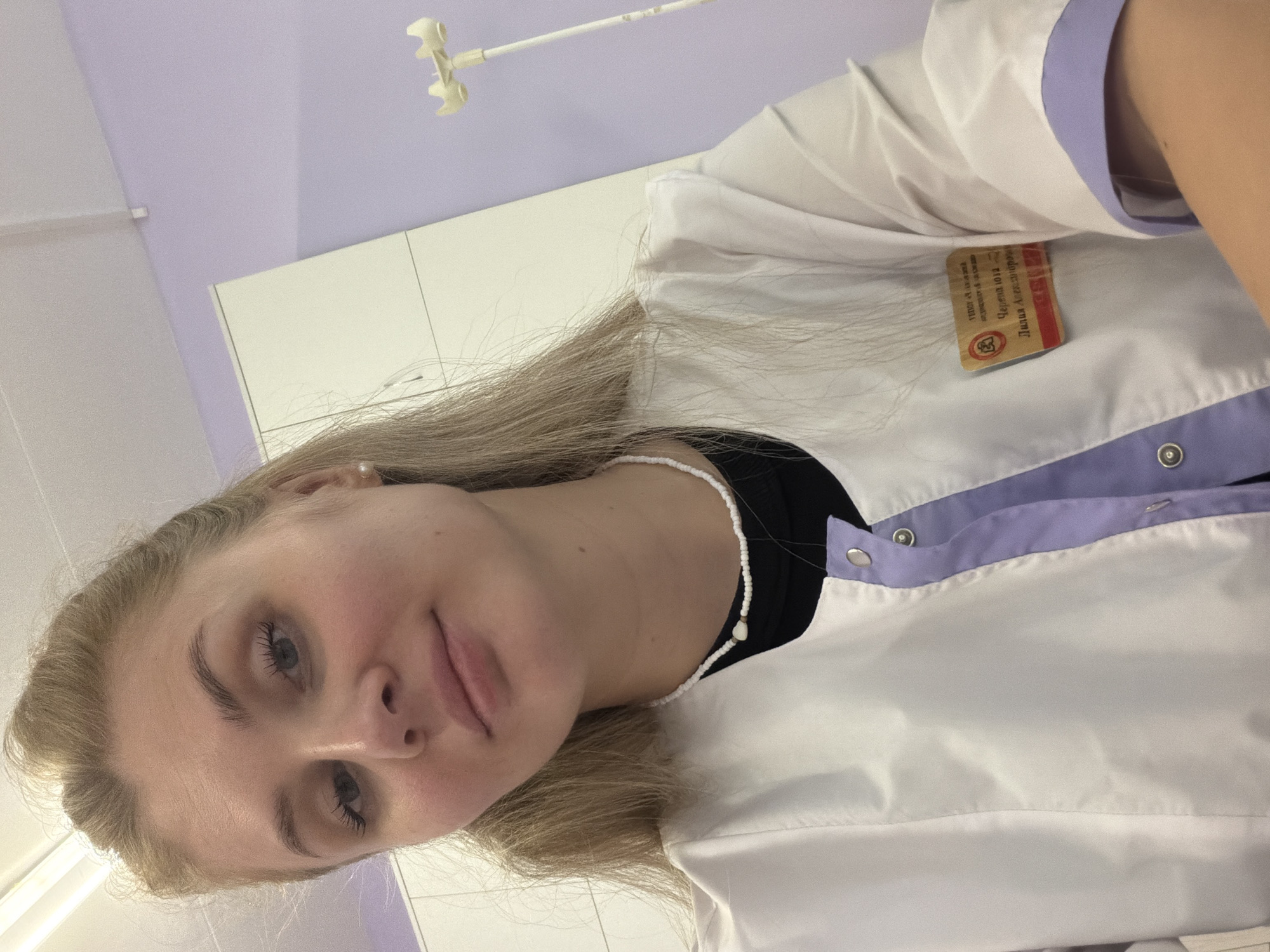 A young adult female, identified by a name badge as Yulia Lidiya Alexandrovna Chernova of Chaikovsky City Hospital (ГБУЗ ПК Чайковская ГБ) in Chaikovsky, Perm Krai, Russia, is captured in a close-up, upward-angled shot. She has long, straight blonde hair, light-colored eyes, and is wearing mascara and a neutral lip color. A small pearl earring is visible in her right ear. She is dressed in a white medical-style uniform top featuring light purple accents on the placket and cuffs, fastened with silver snap buttons. A black long-sleeved shirt is worn beneath the uniform, and a white beaded necklace with a central pearl adorns her neck. The background consists of a light purple wall on the right, a white cabinet or wall on the left, and a white ceiling with a fluorescent light fixture. A medical intravenous (IV) pole is partially visible on the far right. The subject looks directly into the camera with a neutral expression.