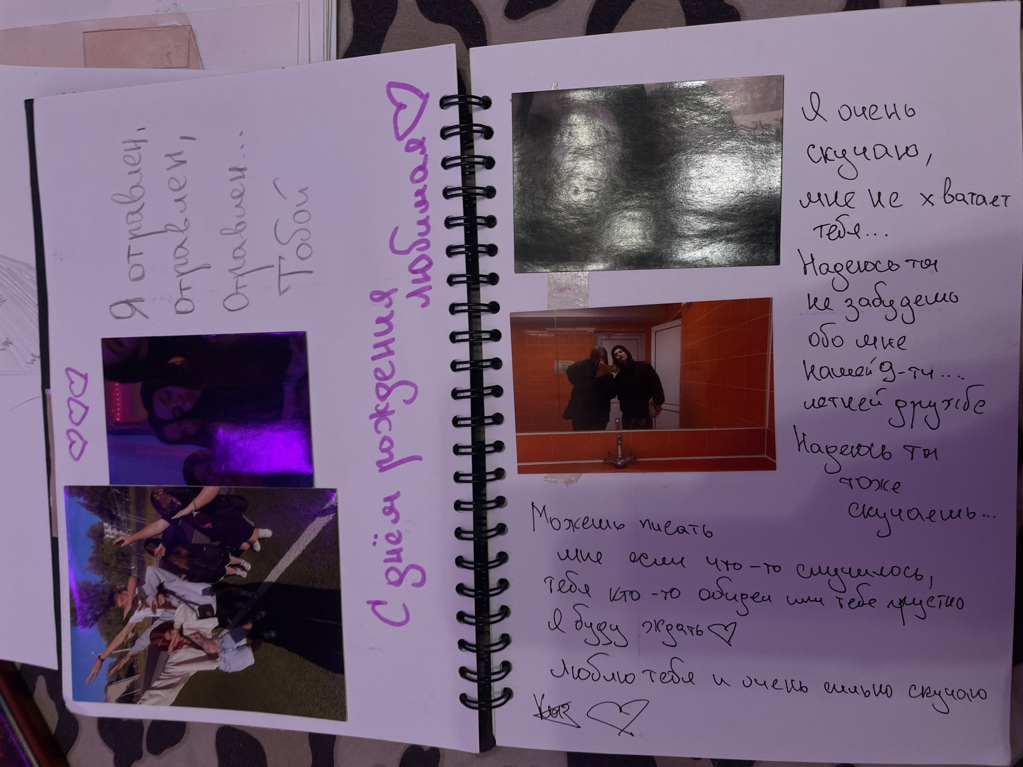The image displays an open spiral-bound notebook with handwritten text and photographs. The notebook is laid flat on a surface with a camouflage pattern. On the right page, there are three pink heart outlines at the top, followed by handwritten Russian text in grey ink. The text reads: "Я отравлен, отравлен, Отравлен... Тобой" which translates to "I am poisoned, poisoned, Poisoned... By you." Below this text is a photograph of a person, possibly indoors.
The left page of the notebook features a large photograph on the top left showing a group of young people posing outdoors, with one person appearing to be doing a "dab" pose. To the right of this photograph is a smaller, vertically oriented photograph with a purple glow, showing two individuals. Below these photographs, a heading in large purple cursive text reads "С днём рождения любимая ♡" which translates to "Happy birthday my love ♡".
Further down the left page, there is a drawing of a stylized heart with an arrow through it. To the right of this drawing is a column of handwritten Russian text in black ink. The text appears to be a personal message or poem, including phrases like "Можешь писать мне если", "тебя кто-то", "Я буду ждать", "люблю тебя" and "и очень сильно скучаю".
On the right side of the left page, starting from the bottom, there are more handwritten Russian text in black ink, which reads: "Я очень скучаю, мне не x хватает тебя... Надеюсь ты не забудешь обо мне какой-g-ту..." translating to "I miss you very much, I lack you... I hope you don't forget about me, any kind of..." This text is partially obscured by photographs.
There are two photographs on the lower portion of the right page. The left photograph shows two people in a room that appears to be a bathroom with orange tiles. The right photograph is a dark, reflective image, possibly a person's reflection or a highly glossy print.
The location of Ladozhskii Trudposelok, Russia, is not depicted or suggested in the image.