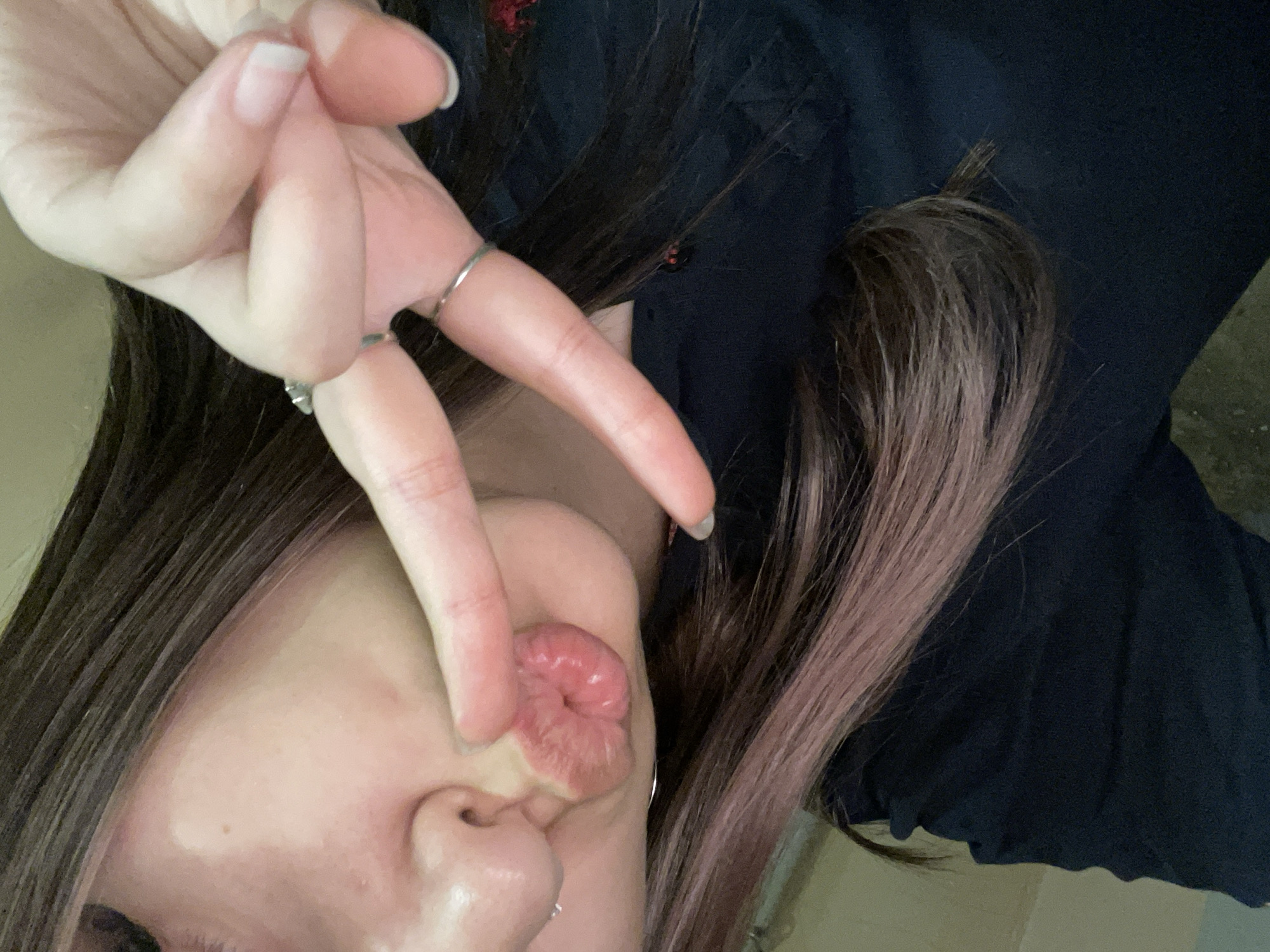 A close-up, rotated portrait shot captures a person, likely female, from the chin upwards, making a "finger heart" gesture with their hand while puckering their lips. The hand is in the foreground, with fingers forming a heart shape, and two silver-toned rings are visible on the fingers. The nails are neatly trimmed and natural. The person's face shows pale skin, a small mole or blemish near the nose, and lips tightly puckered into a "kissing" pose. Dark, possibly brown, hair with subtle lighter highlights, perhaps red or purple, frames the face and falls across the dark clothing visible on the shoulder. The clothing appears to be a dark fabric, possibly black or navy, with some subtle texture. The overall impression is one of playfulness or affection. No specific background or location details are discernible beyond the person and their immediate attire. There is no visible text.