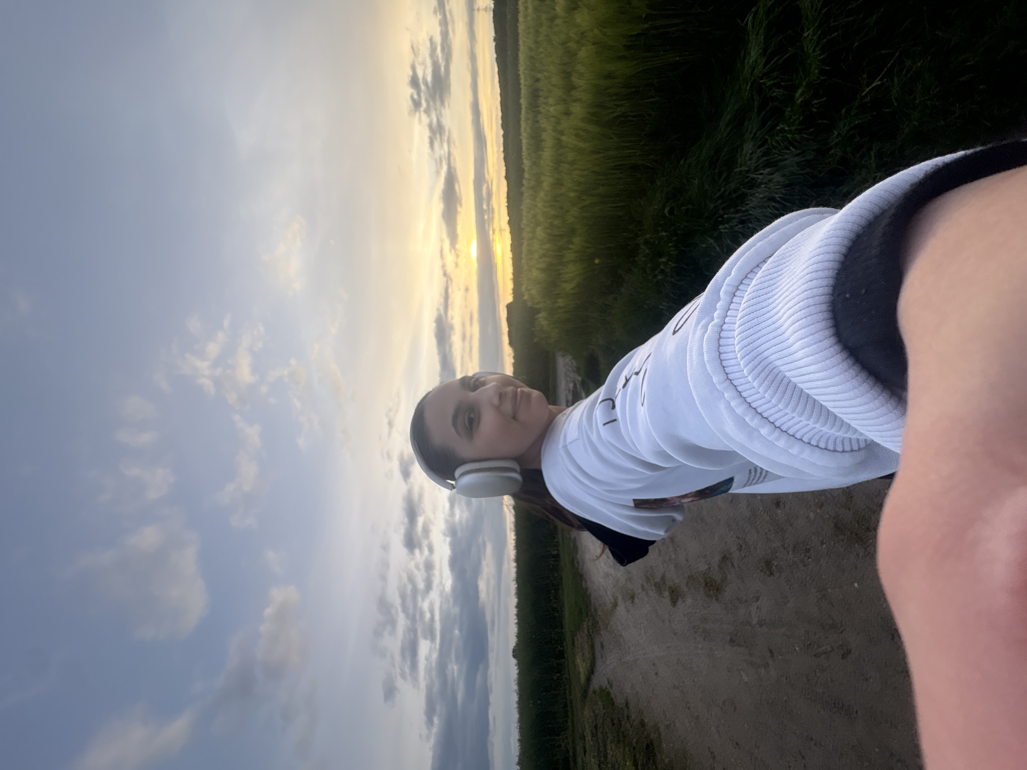 A daytime, outdoor selfie shows a woman in the countryside near Mönchengladbach, Germany. She is wearing white headphones and a white sweatshirt, standing on a dirt path flanked by fields. The sky is visible in the background, displaying a mix of clouds and a setting or rising sun. The photo is taken at an angle.