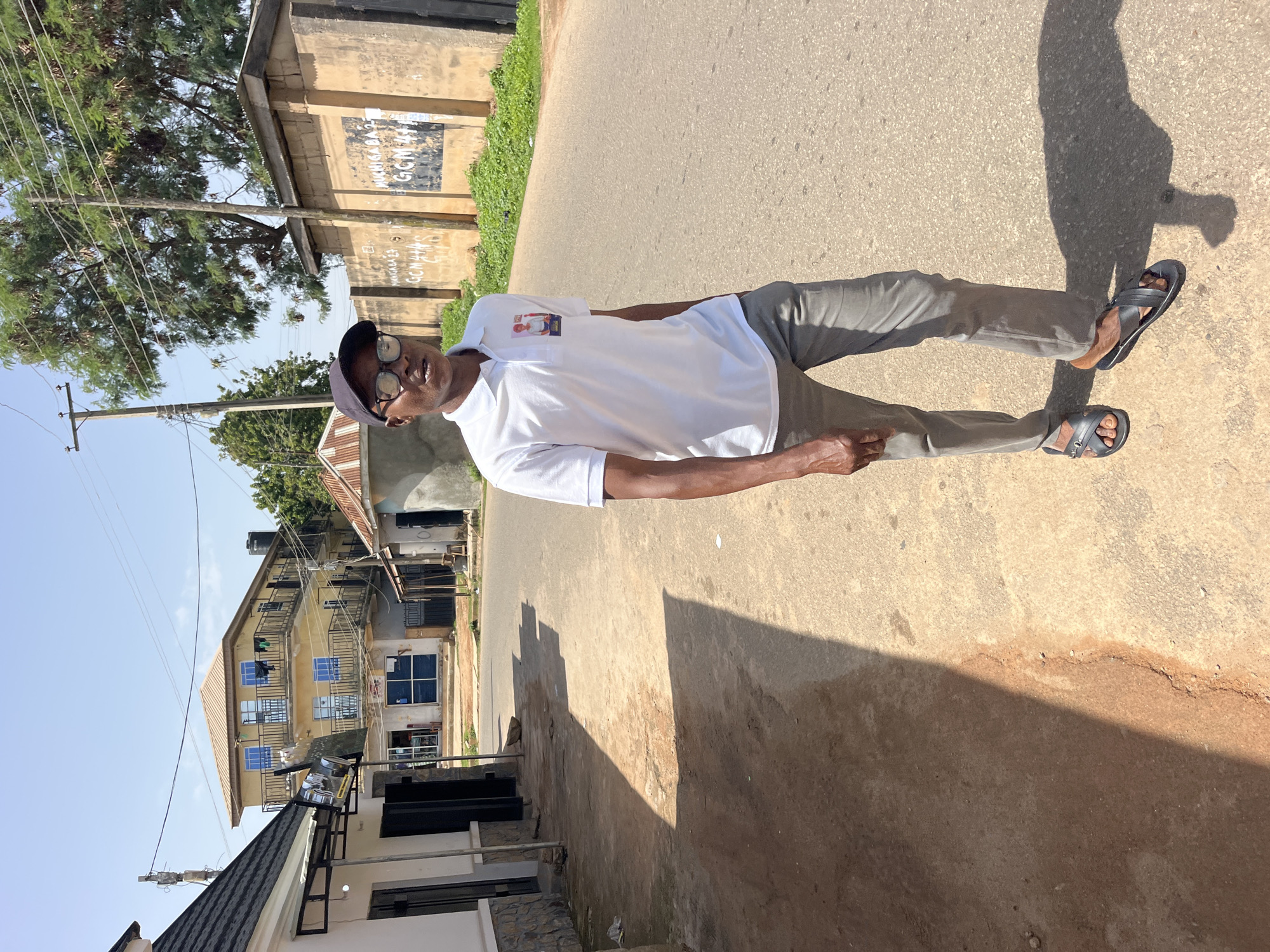 A young Black man stands in the middle of a paved road under a clear blue sky. He is wearing a white polo shirt with a small image on the left chest, dark-rimmed glasses, and a black baseball cap. His pants are grey, and he is wearing black sandals. He is looking slightly to his left and smiling. Behind him, on the right side of the road, is a light-colored concrete wall with some graffiti on it. On the left side of the road, there are several buildings: a white structure with a grey stone base, a yellow building with multiple stories and balconies, and a smaller, older-looking structure with a corrugated metal roof. Trees and utility poles are visible in the background. The road is an asphalt surface, and there is a significant shadow cast on the left side of the road.