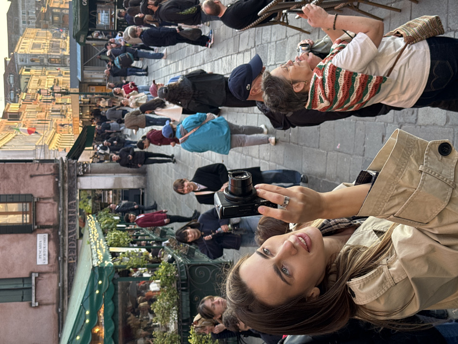 The image captures a bustling pedestrian street in Venice, Italy, specifically identified by a sign reading "PESCARIA S.BORTOLOMEO," indicating the area near Campo San Bartolomeo. In the left foreground, a young woman with long, light brown hair wears a beige trench coat over a plaid shirt and holds a black compact digital camera with both hands, aiming it slightly upward and to the left. She has a silver-toned ring on her right ring finger and a faint smile. To her right, in the mid-ground, an older woman with short grey hair and glasses wears a white t-shirt, blue jeans, and a red, green, and white striped knitted shawl; she holds a smartphone, actively aiming it forward as if taking a photograph. A man in a dark blue baseball cap is partially visible behind her.
The left side of the street features an outdoor cafe with dark green awnings adorned with string lights, below a building with a faded pink facade and windows with closed shutters. Another sign on this building reads "PEDAVENA." Several individuals are seated at the cafe's tables. The stone-paved street is crowded with numerous pedestrians of varying ages and attire, walking in multiple directions. In the mid-ground, behind the foreground subjects, a woman in a dark coat holds a retro-style compact camera. The background reveals additional Venetian buildings with traditional architecture, showcasing faded yellow and orange facades under warm, late afternoon sunlight. An Italian flag is visible on a pole attached to a building in the distant background.
