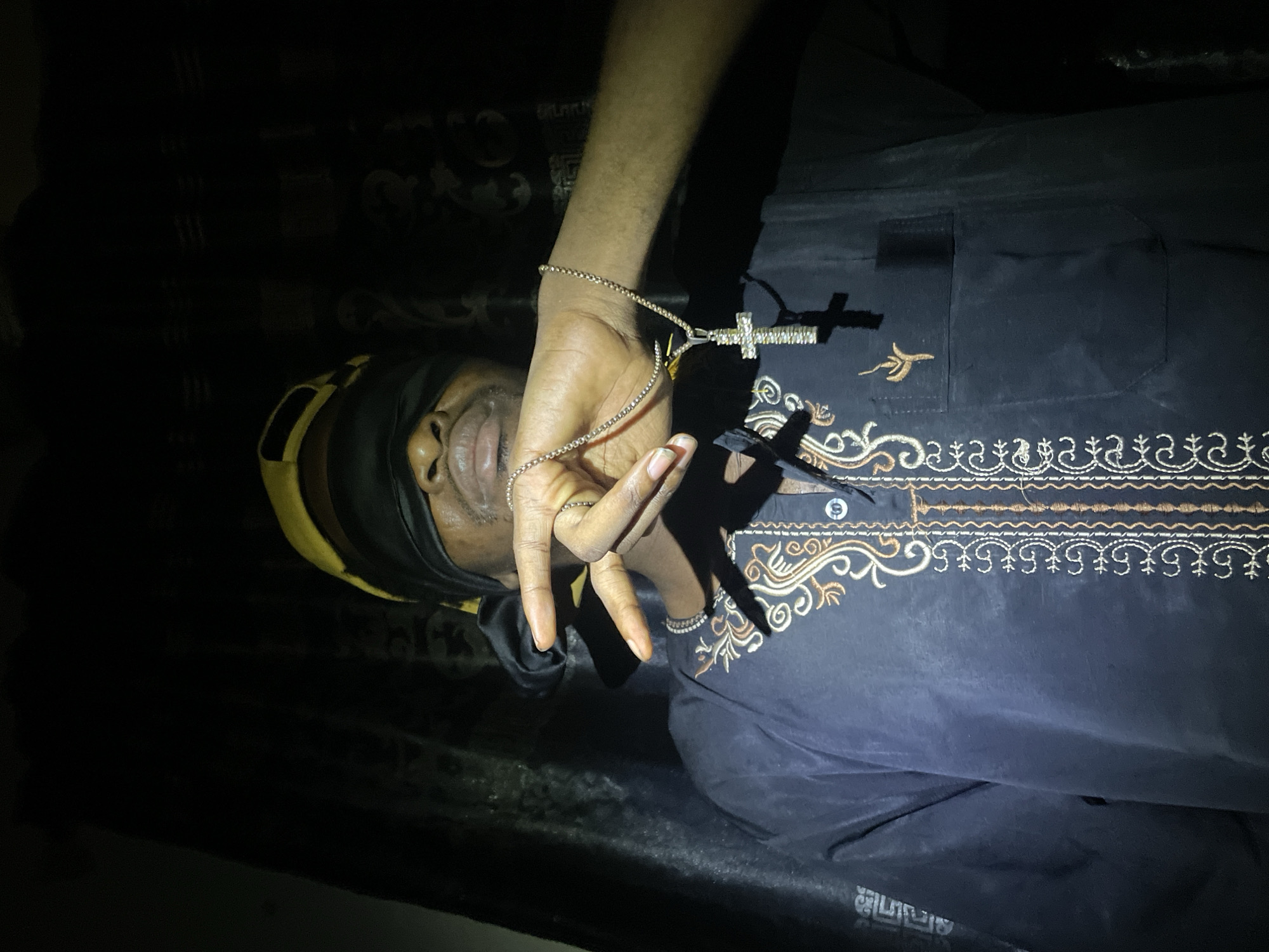 An African male lies on his back in a dimly lit room, his eyes covered by a black blindfold with yellow trim. He wears a black shirt with intricate gold and white embroidery along the chest and collar. In his right hand, he holds a silver chain necklace with a cross pendant. He also wears rings on his fingers. The location is unknown and there is no explicit or implicit evidence that this image was taken in Lokoja, Nigeria.